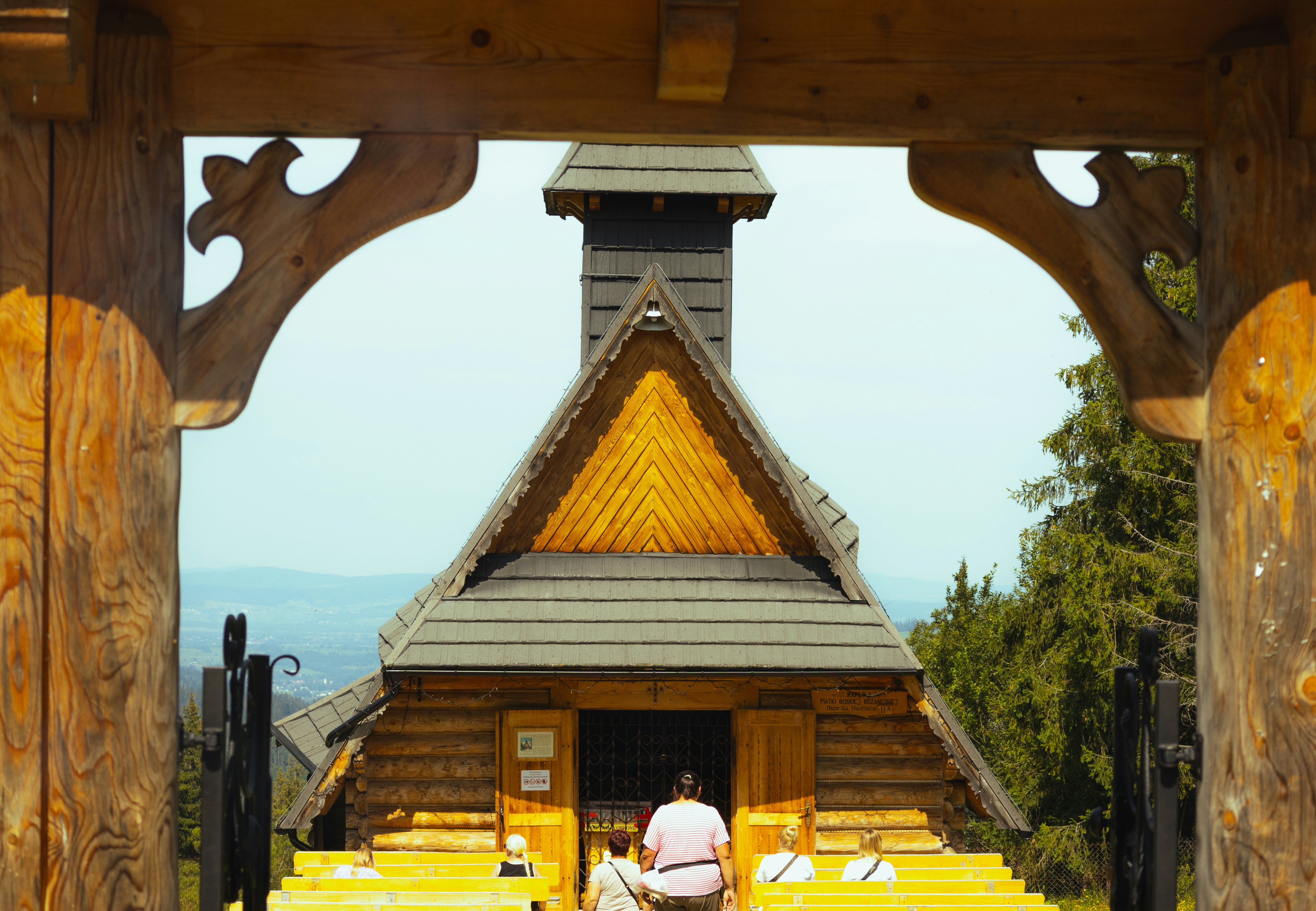 People gather at a rustic wooden chapel with yellow benches.