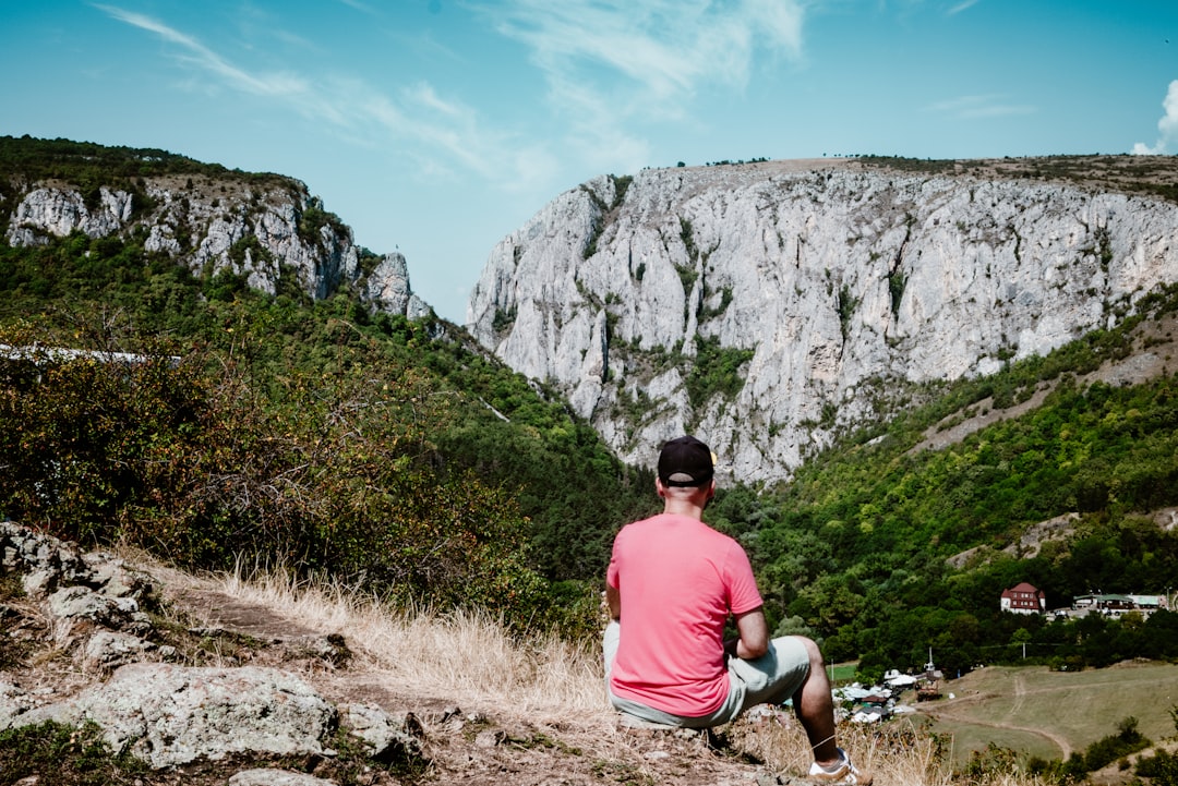 A man sitting on top of a hill next to a lush green hillside, A serene moment as an individual admires the majestic clif