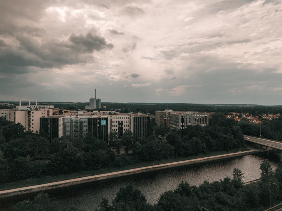 The image presents an aerial view of a cityscape, with a river flowing through it and several buildings lining its banks. The sky above is filled with clouds, casting a soft light over the scene. In the foreground, there are trees that add a touch of nature to the urban setting. The perspective of the photo gives a sense of depth and distance, making the city appear vast and expansive.