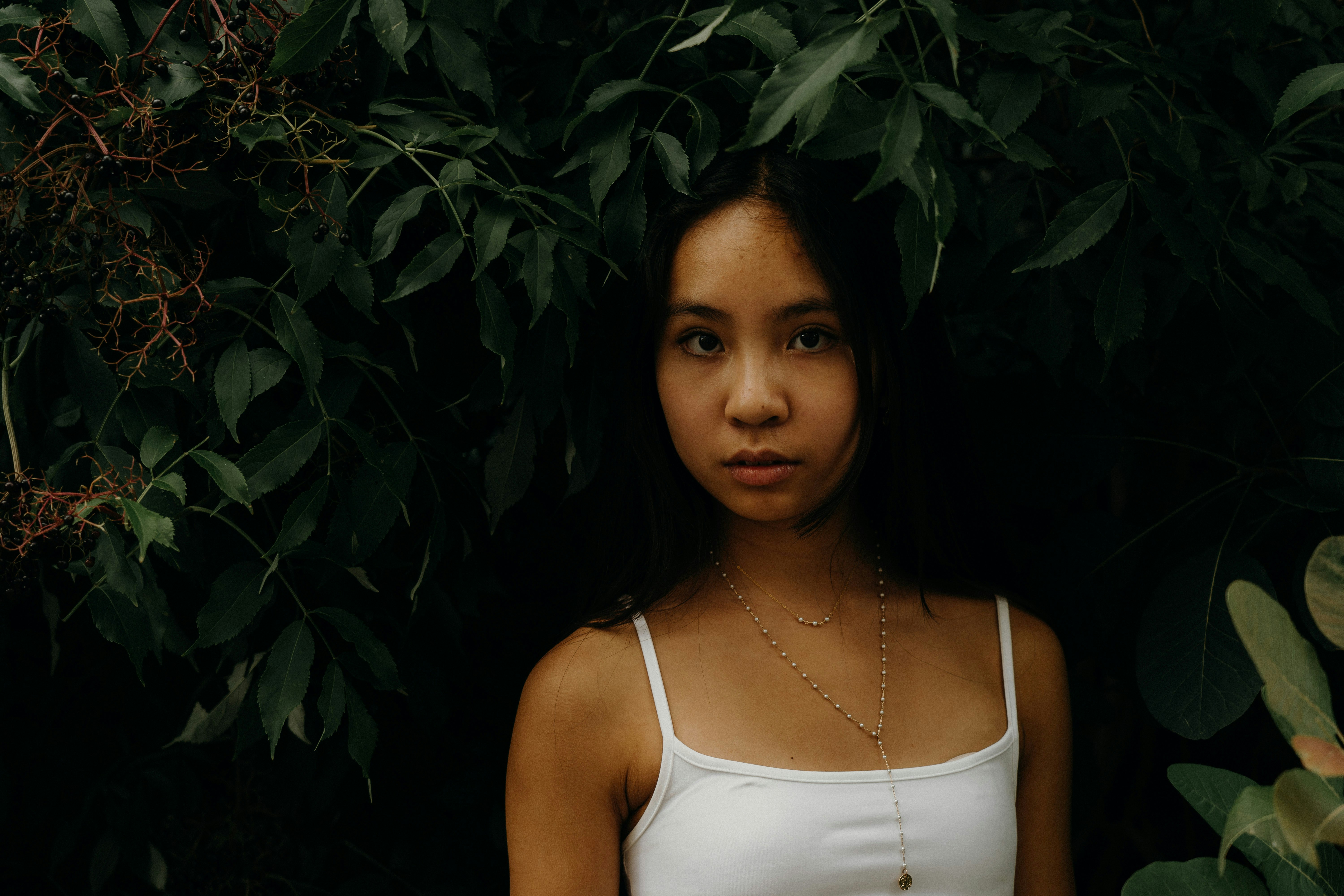 A woman standing in front of a bush wearing a white tank top