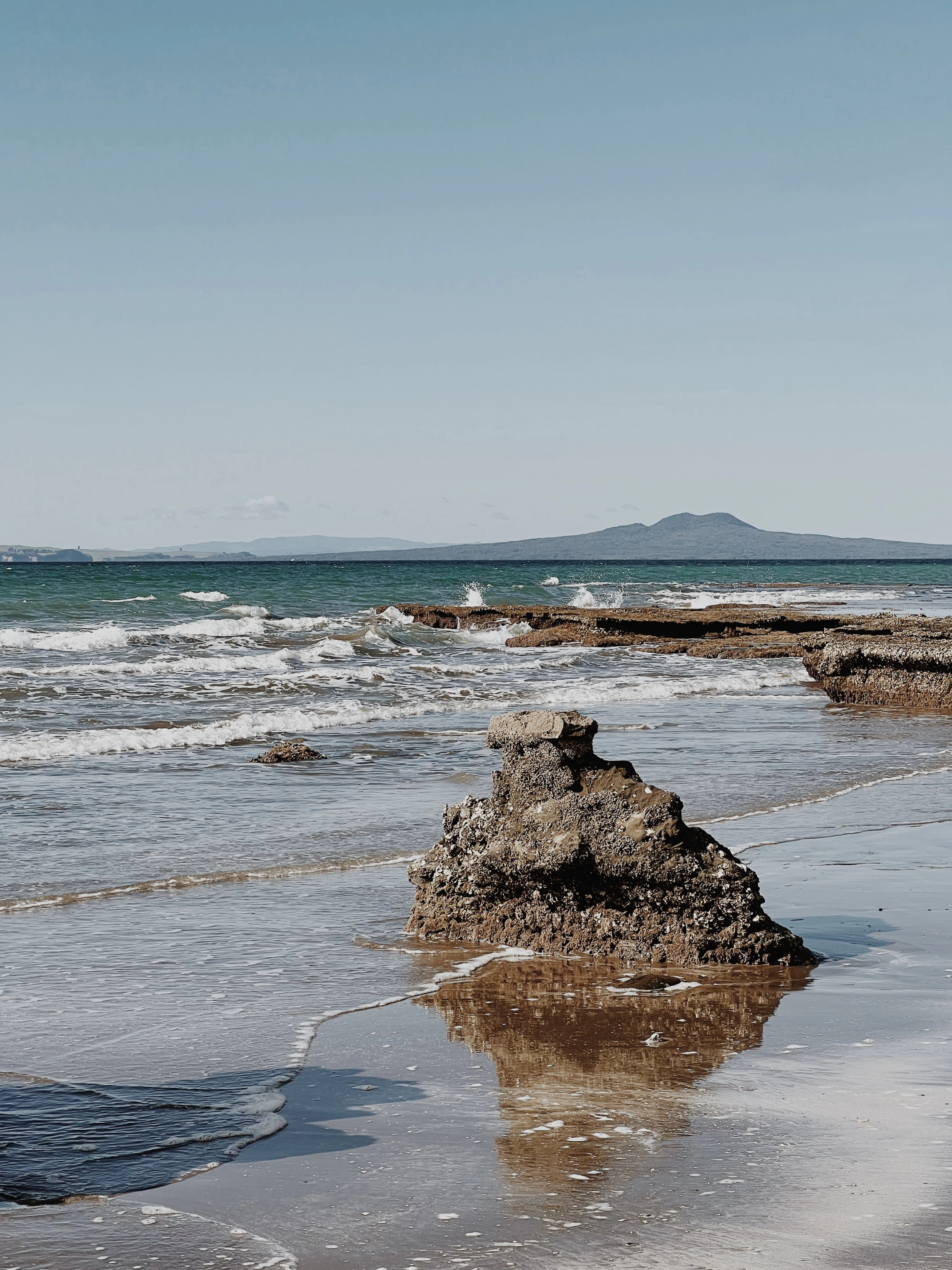A weathered volcanic rock on a beach with rolling waves and a distant island on the horizon.