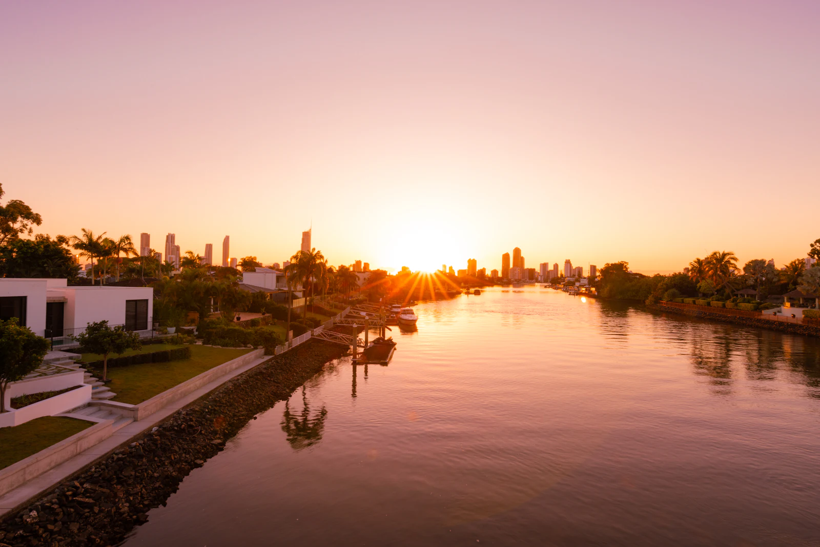 Low waterfront houses and palms silhouetted against a sunset over calm water