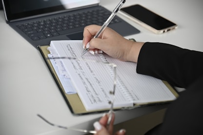 A person writing on a notebook with a laptop in the background