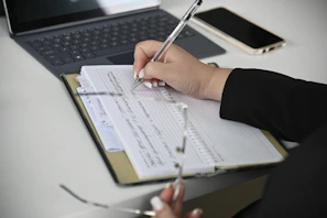 A person writing on a notebook with a laptop in the background