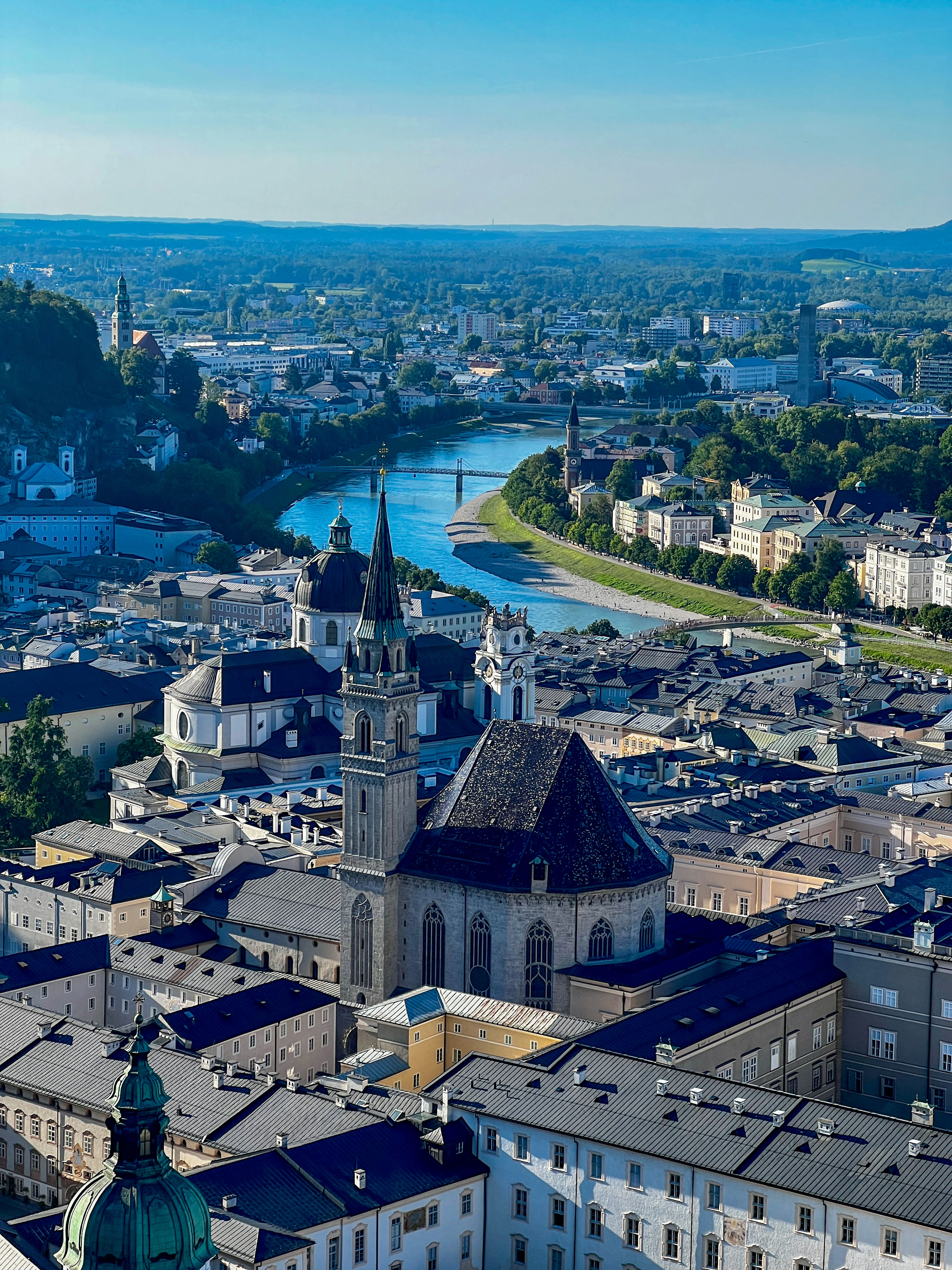 Aerial view of Salzburg's rooftops with the Salzach river curving through the city under a clear blue sky.
