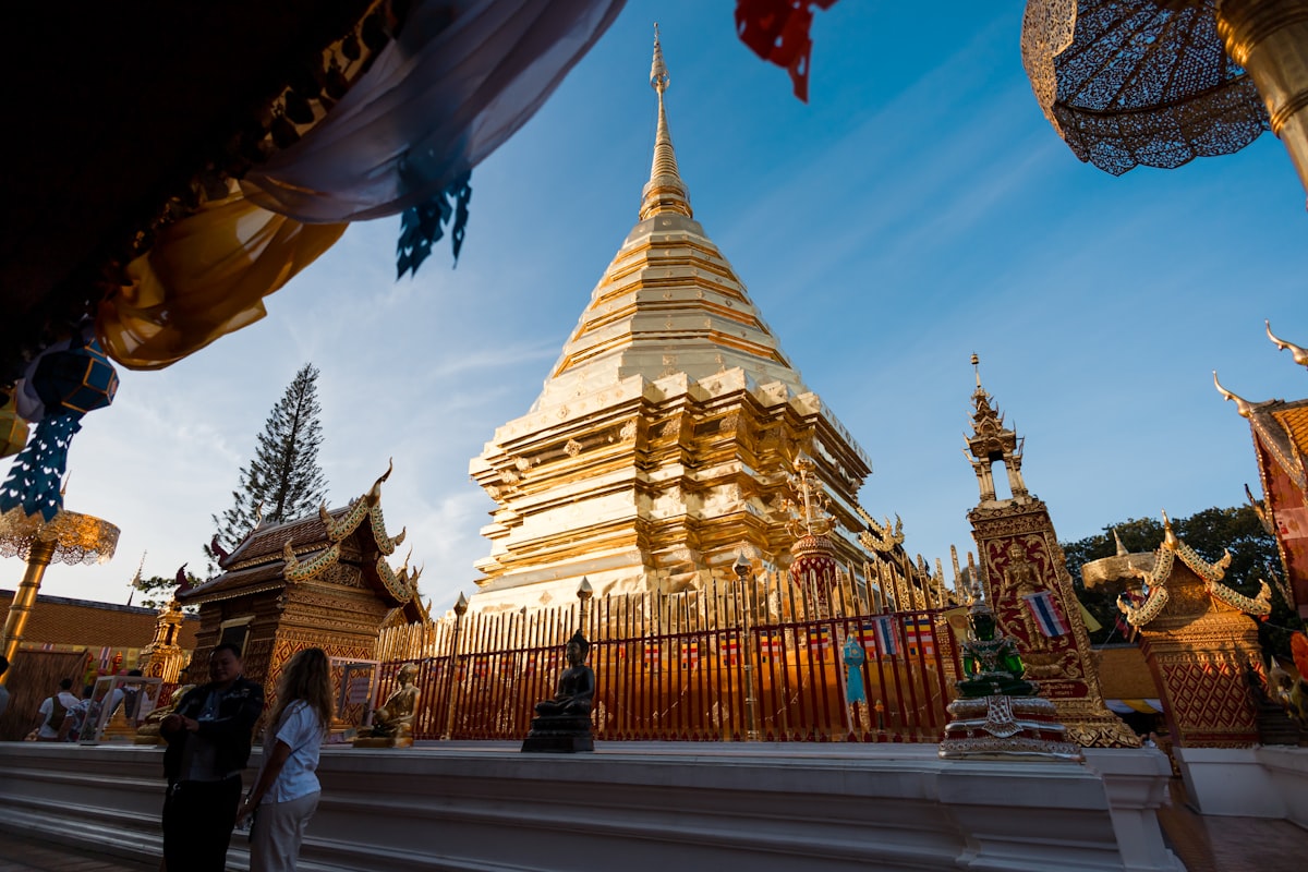 A golden chedi seen through an ornate temple gateway in Chiang Mai, a few visitors at its base and blue sky with clouds