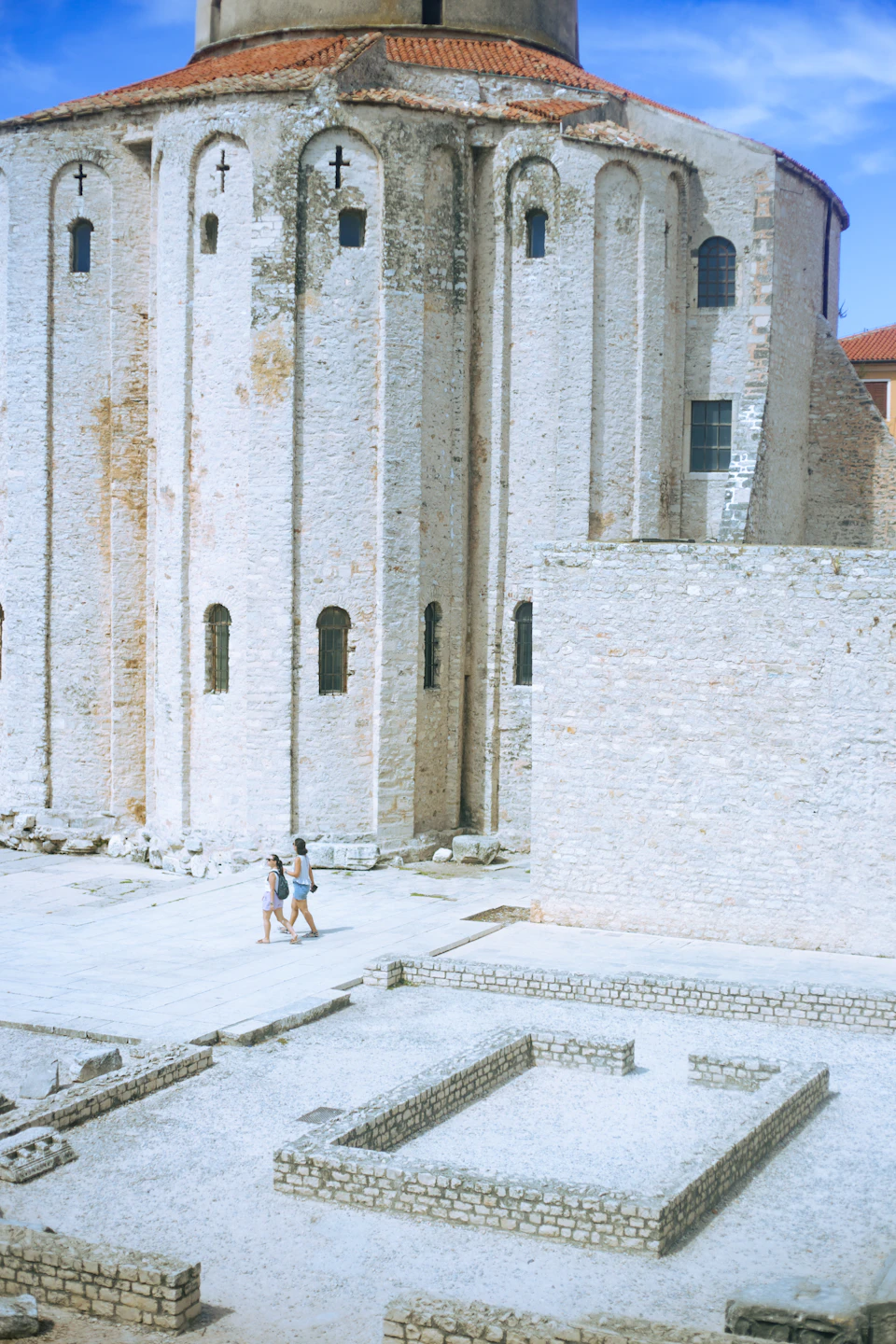 The image depicts a large, white stone building with multiple arches and windows, standing tall against a clear blue sky. The building is situated within an open courtyard, which features a few scattered rocks and boulders. Two individuals can be seen walking in front of the building, adding a sense of scale to the scene.

The image does not contain any discernible text or countable objects, but it captures the architectural beauty of the stone structure and the peaceful atmosphere of the courtyard.