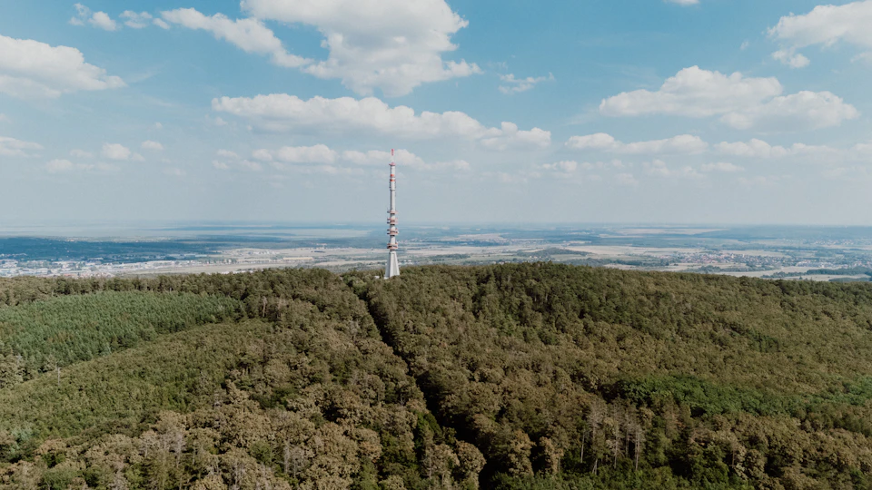 The image presents an aerial view of a large, tall tower with multiple antennas and a pointed top, standing prominently against a backdrop of lush green trees and rolling hills. The tower is situated on a hilltop, surrounded by a dense forest that extends into the distance. The sky above is a clear blue, dotted with fluffy white clouds.
