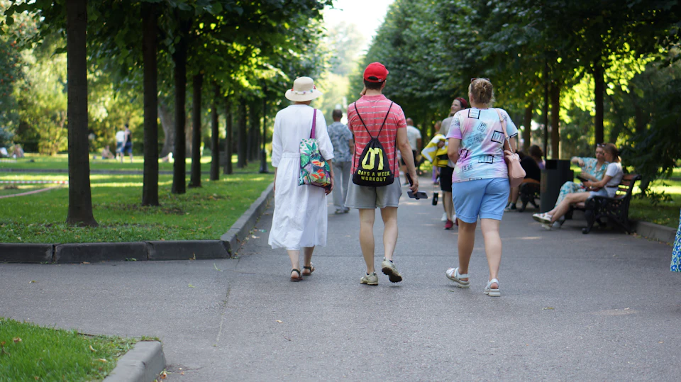 The image depicts a lively park scene with several people walking along a tree-lined path. The individuals are carrying various bags, including backpacks and handbags, suggesting they may be tourists or travelers. Some of them appear to be wearing hats for sun protection. In the background, there is a bench where one person can be seen sitting, possibly enjoying the park's atmosphere.

The trees lining the path provide shade and add to the overall ambiance of the scene. The image captures the essence of a typical day in a park, with people going about their daily activities while taking in the beauty of nature.