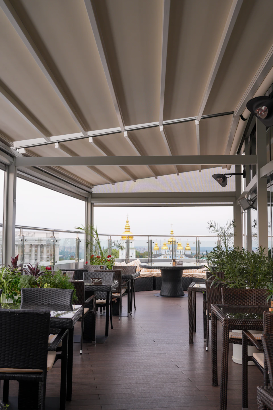 The image depicts a rooftop patio with a large white canopy providing shade over several tables and chairs arranged on wooden floors. The patio is adorned with potted plants, adding greenery to the space. A few people can be seen seated at the tables under the canopy, enjoying their time in this outdoor dining area.

In the background, there are buildings visible through a glass wall that adds depth to the scene and enhances the sense of openness. The overall atmosphere is one of relaxation and leisure, with the patio serving as an inviting space for people to gather and enjoy each other's company.