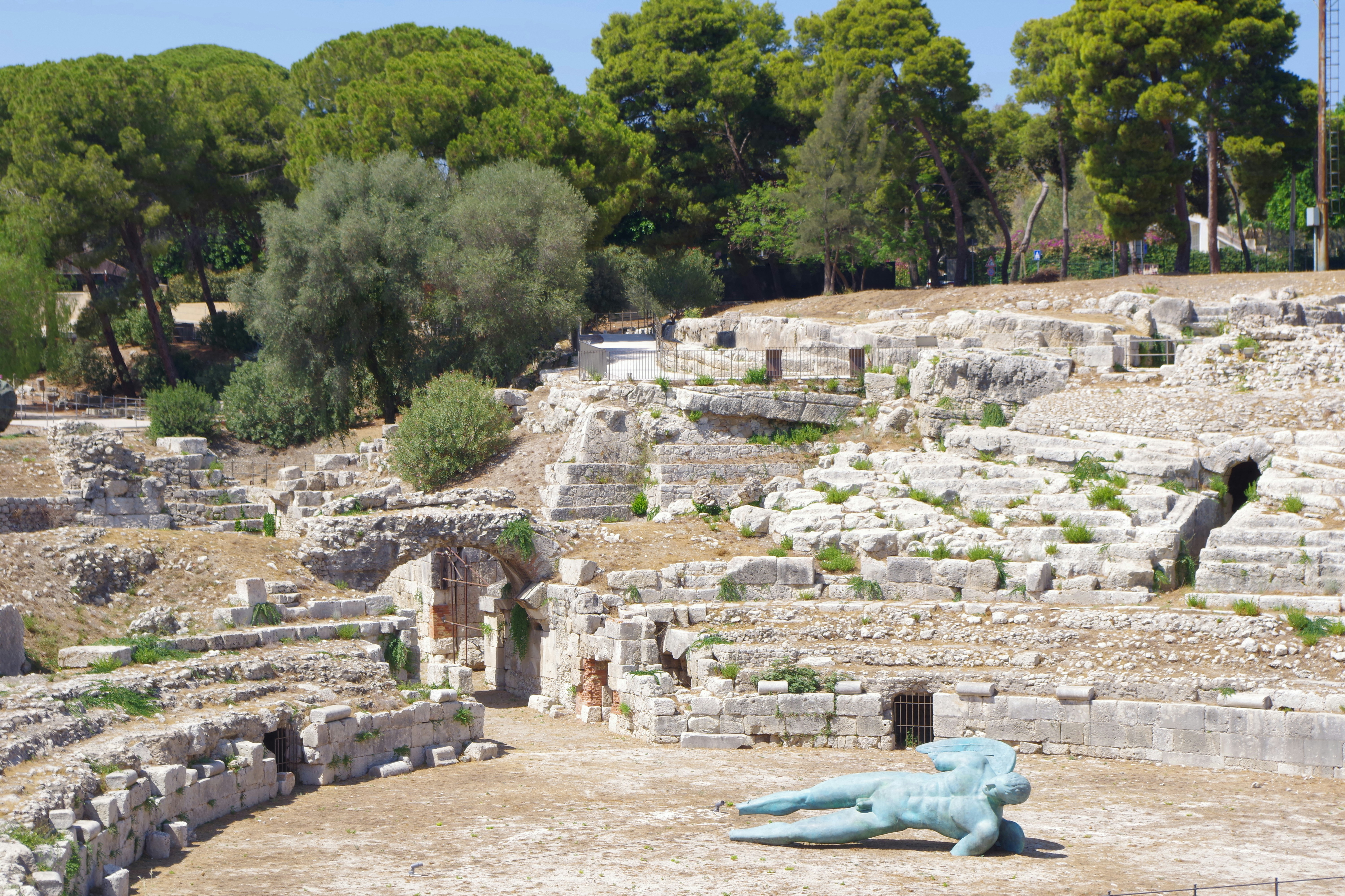 Blue reclining sculpture rests among sunlit ancient amphitheater ruins.