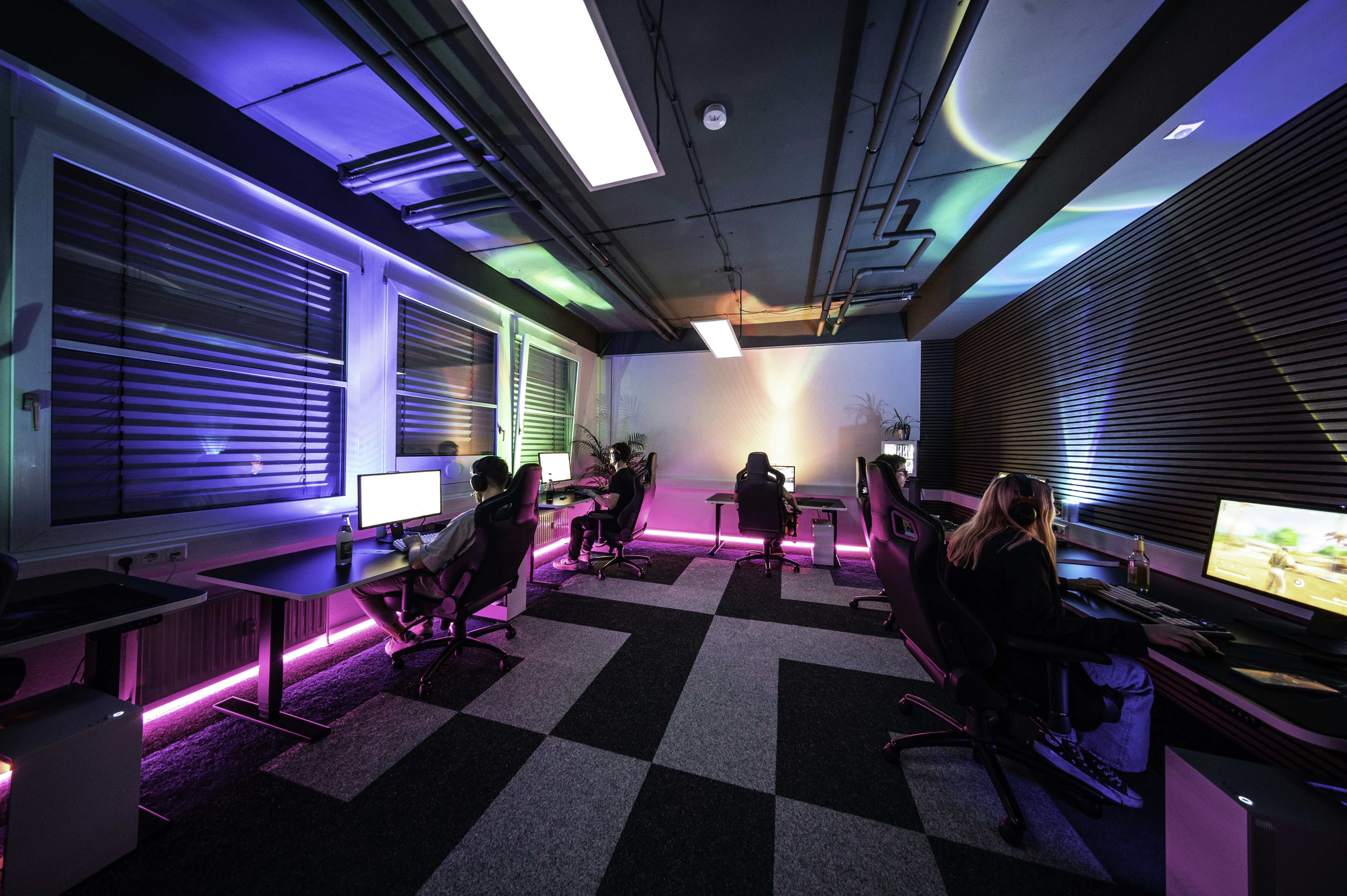 A group of people sitting at desks in a room, 1337Camp offers a unique environment perfectly tailored to the needs of gamers and creatives. Our facility features customizable lighting and LED floor lights that change colors to create the ideal atmosphere. The dojo-like design of the camp promotes a balance of relaxation and focus, allowing you to actively unwind while staying driven towards your goals. At 1337Camp, technology and design come together to create an inspiring and productive space that empowers you to reach your full potential.