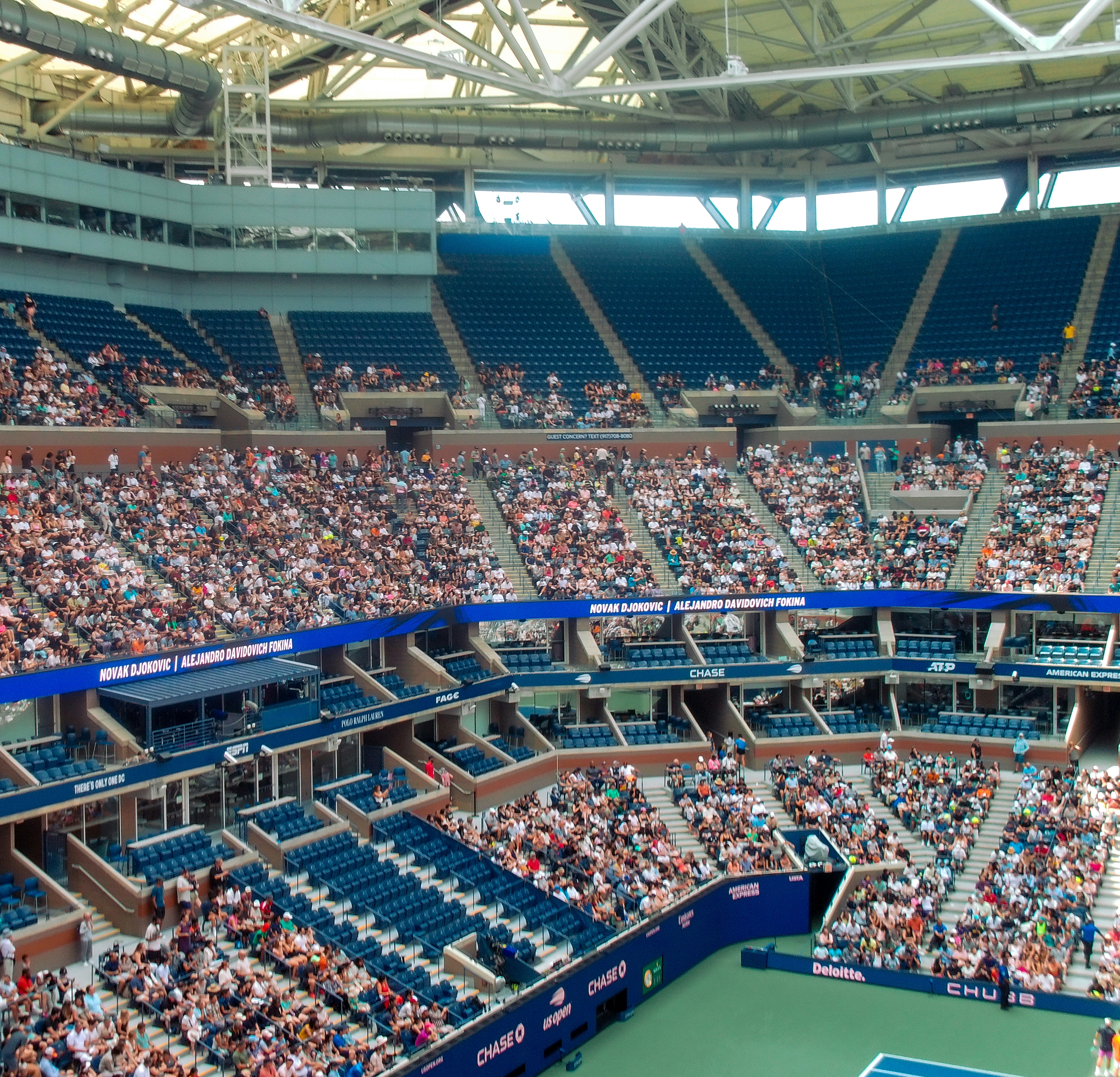 Pristine blue hard court tennis facility with stadium seating viewed from above in bright daylight