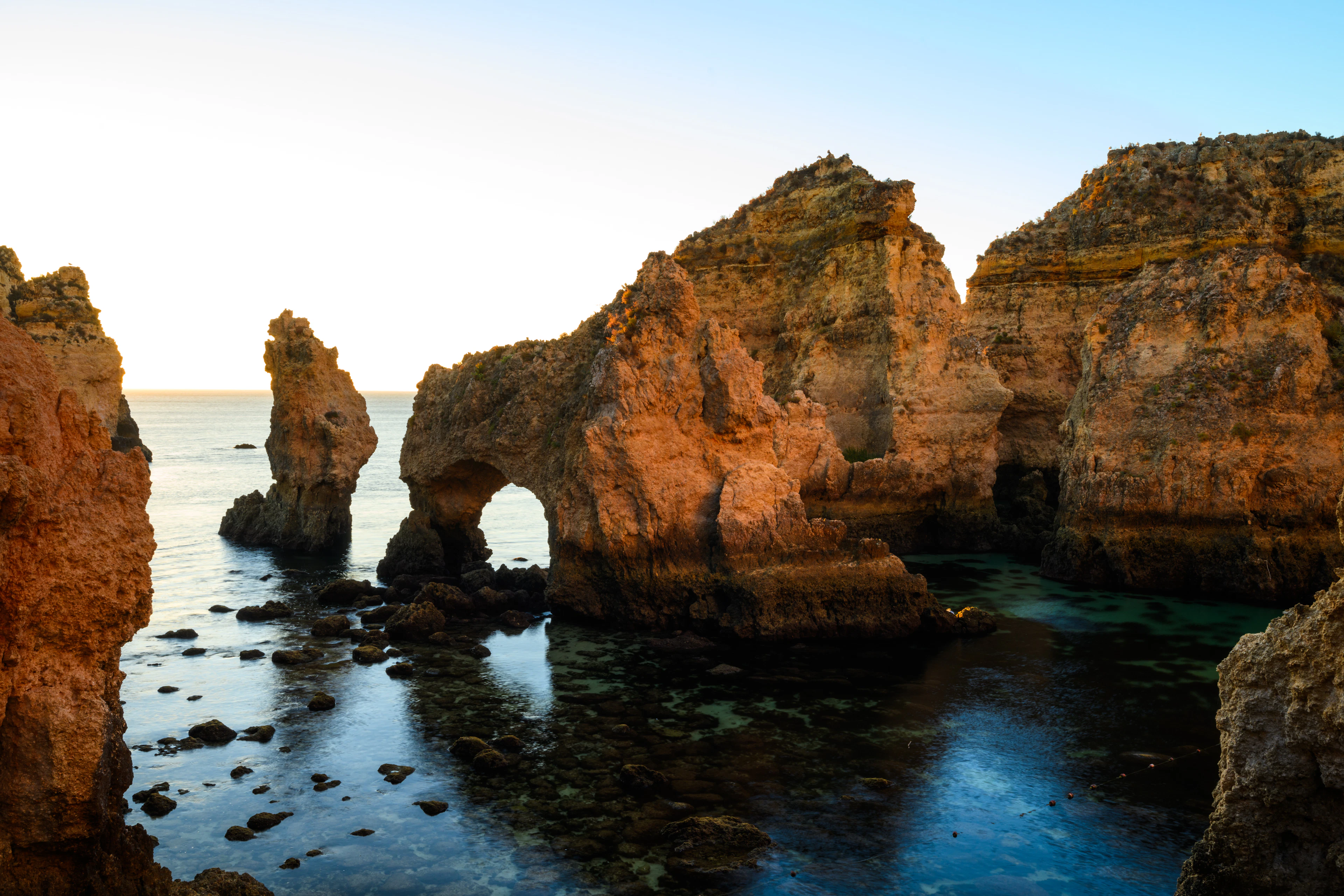 Low golden light striking the ochre sea stacks and natural arch of Ponta da Piedade near Lagos, with clear green water pooling over dark rock in the foreground.
