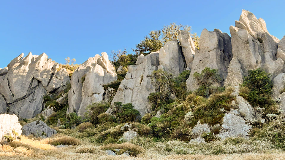 The image depicts a rugged, rocky cliff face with numerous large, jagged rocks and boulders. The cliff is covered in a mix of green shrubs and grasses that add color to the otherwise monochromatic landscape. The sky above is a clear blue, providing a striking contrast to the earthy tones below. The perspective of the image is from ground level, looking up at the imposing height of the cliff face.