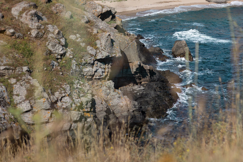The image depicts a rocky cliff overlooking a serene beach scene. The cliff is covered in various shades of gray and brown, with patches of green vegetation peeking through. A few small rocks are scattered around the base of the cliff. Beyond the cliff, the ocean stretches out to meet the sky at the horizon line. The water appears calm, reflecting the clear blue sky above.