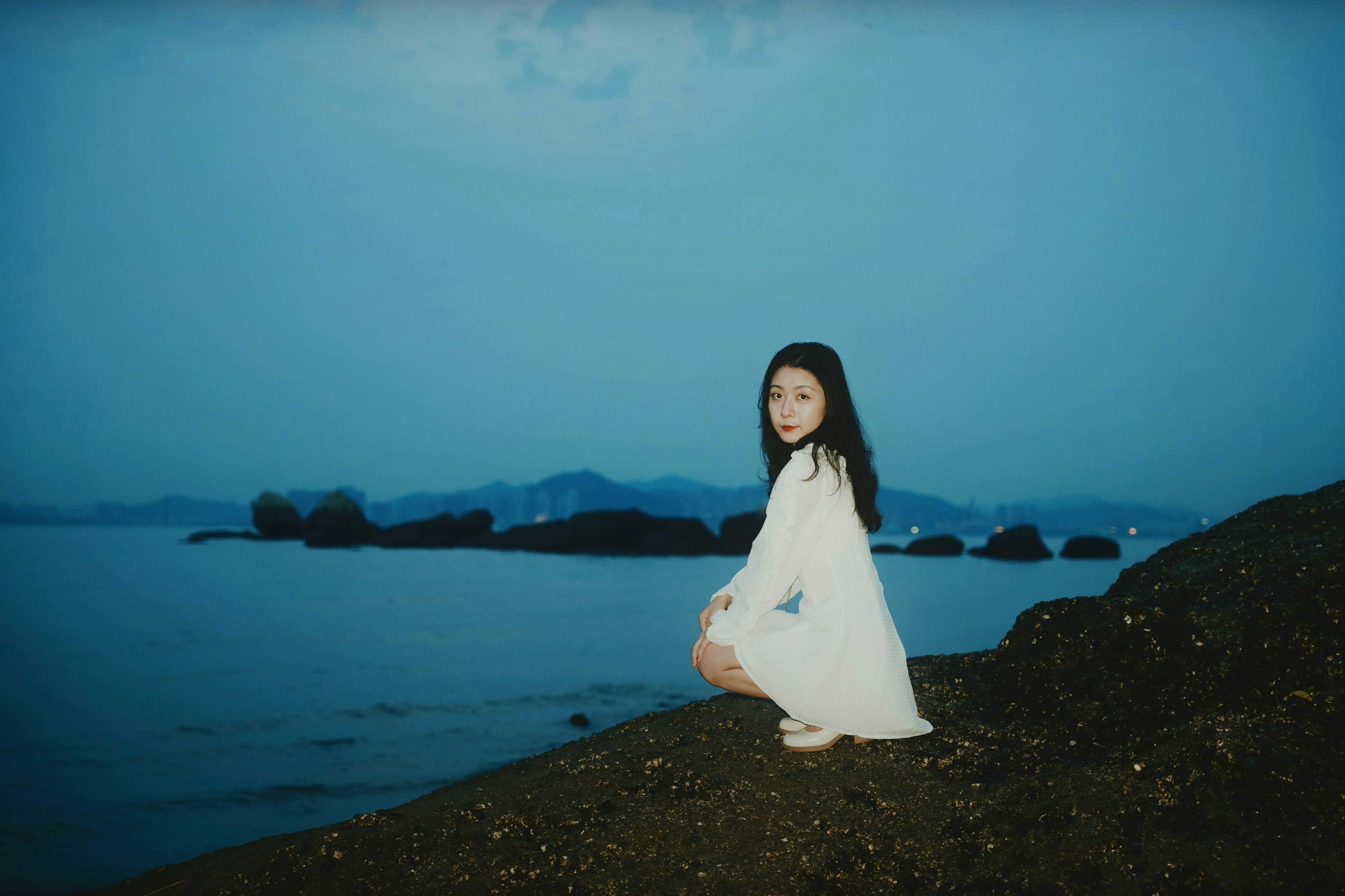 A woman in a white dress sitting on a rock by the water
