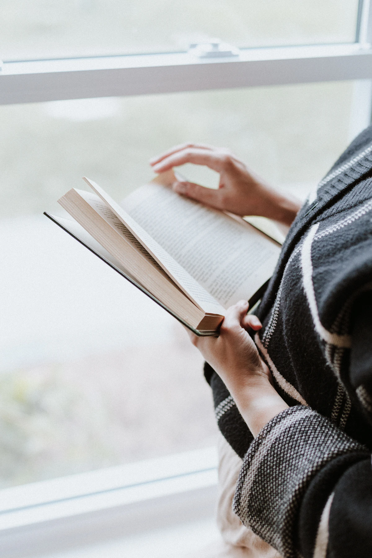 A person reading a book by a window, soft daylight illuminating the pages.