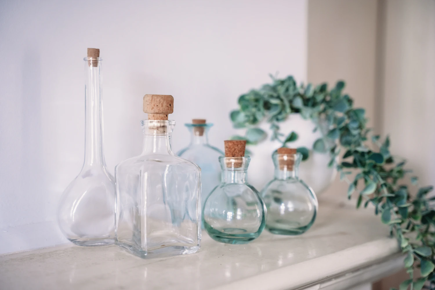 Clear corked apothecary bottles of different shapes on a white mantelpiece beside trailing eucalyptus