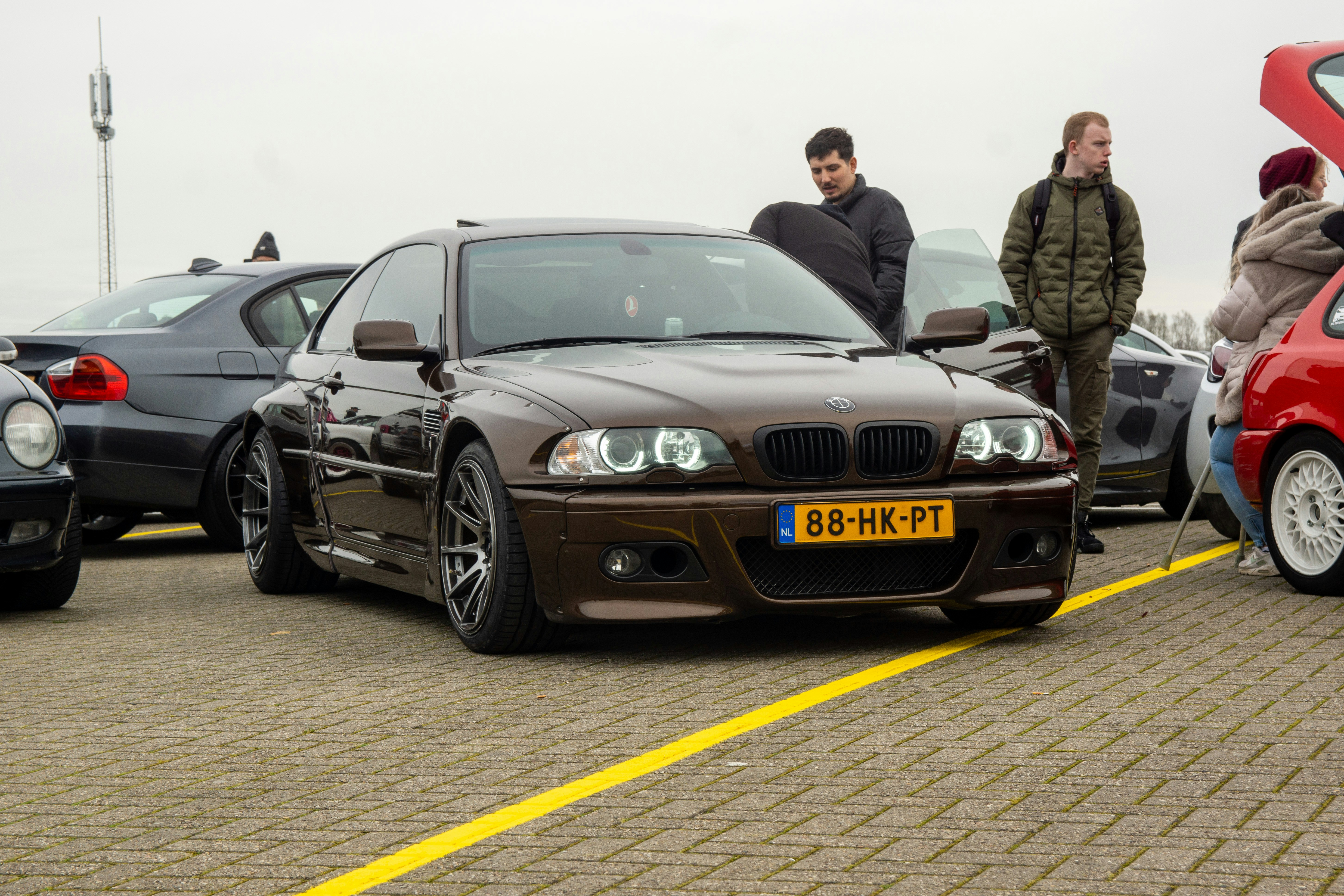 Car enthusiasts gather around a custom brown BMW coupé outdoors.
