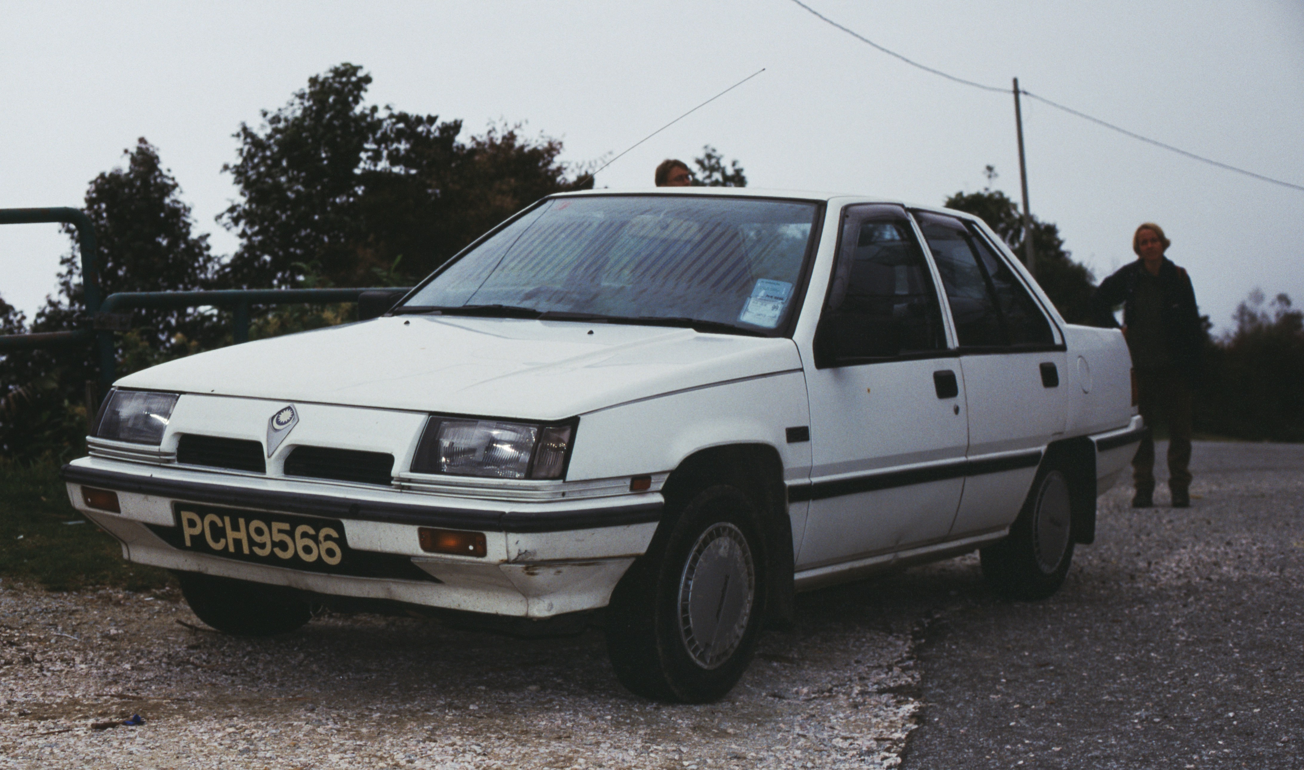 The image shows a white malaysian Proton Saga parked on a gravel road, with trees in the background. The car has a vintage design, likely from the 1980s or early 1990s, and bears a license plate number 