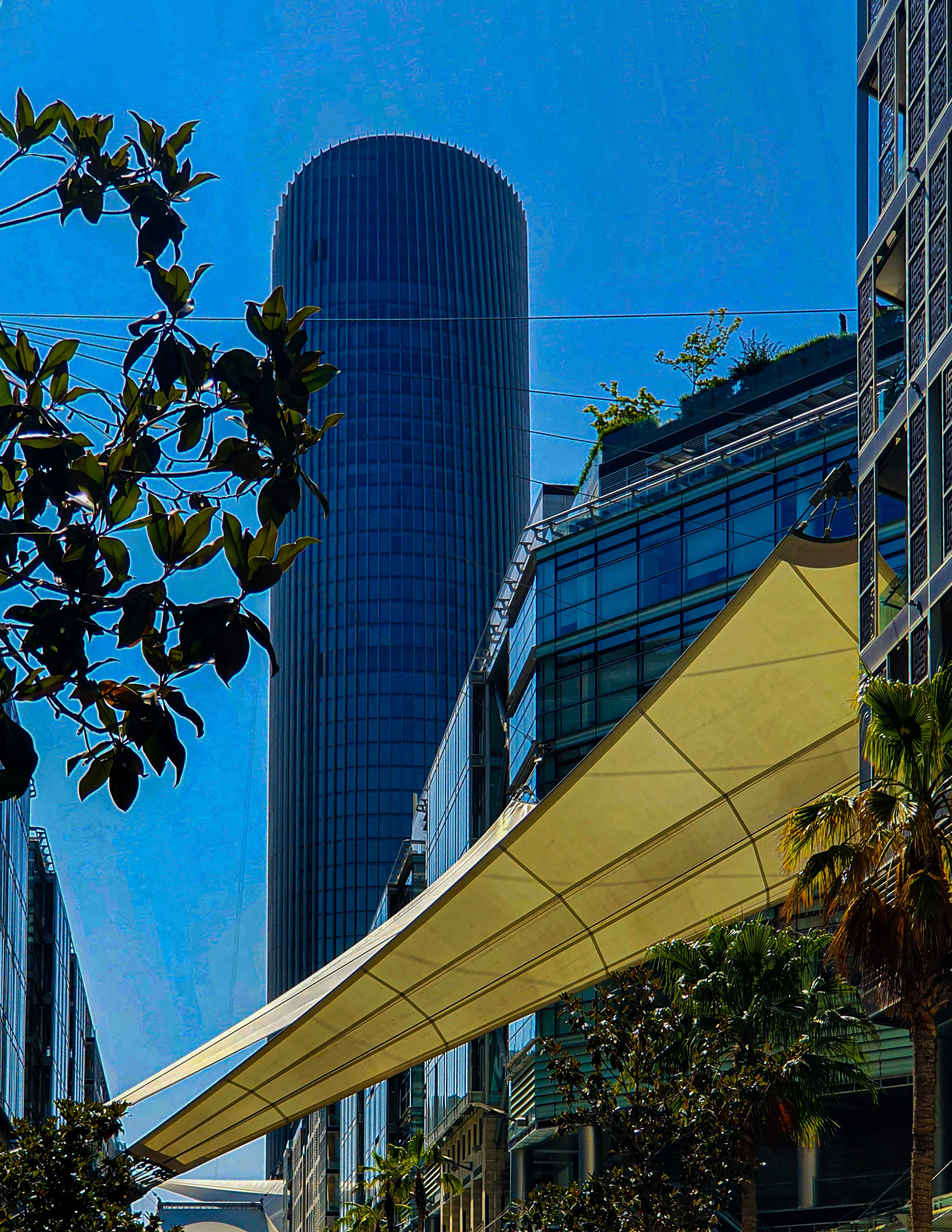 A curved glass skyscraper rises above a fabric shade sail and greenery under a deep blue sky in Amman.