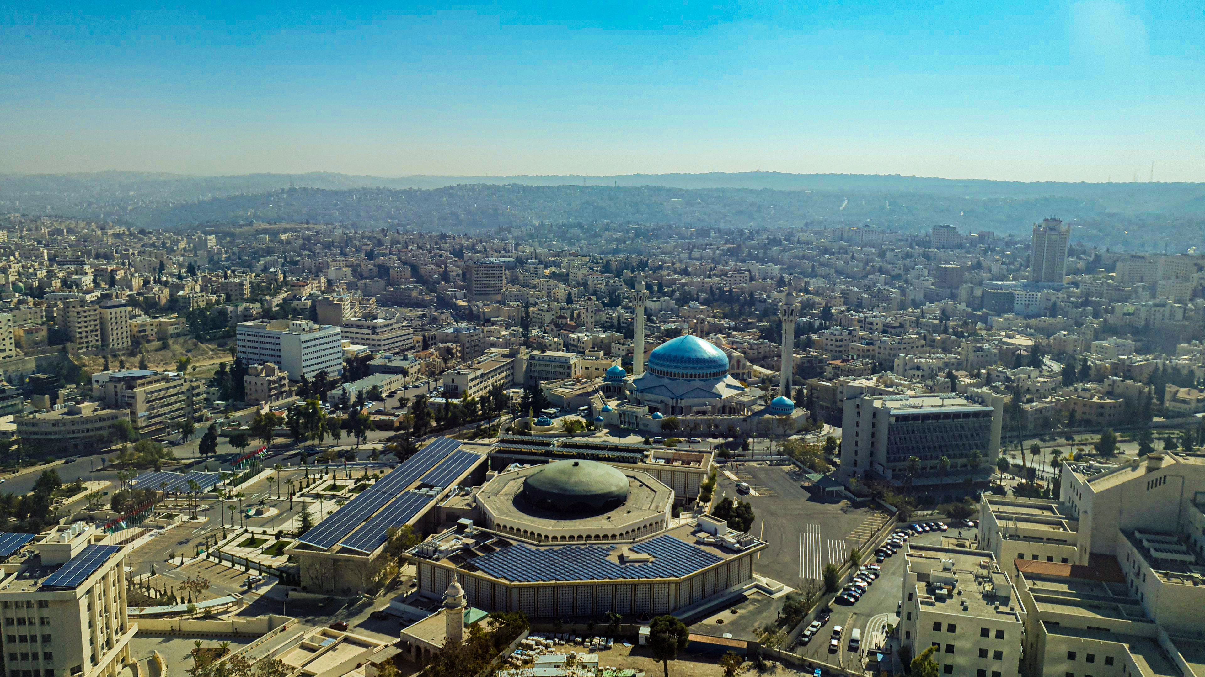 Aerial view of Amman's dense hillside cityscape centered on the blue-domed King Abdullah I Mosque under a hazy sun.