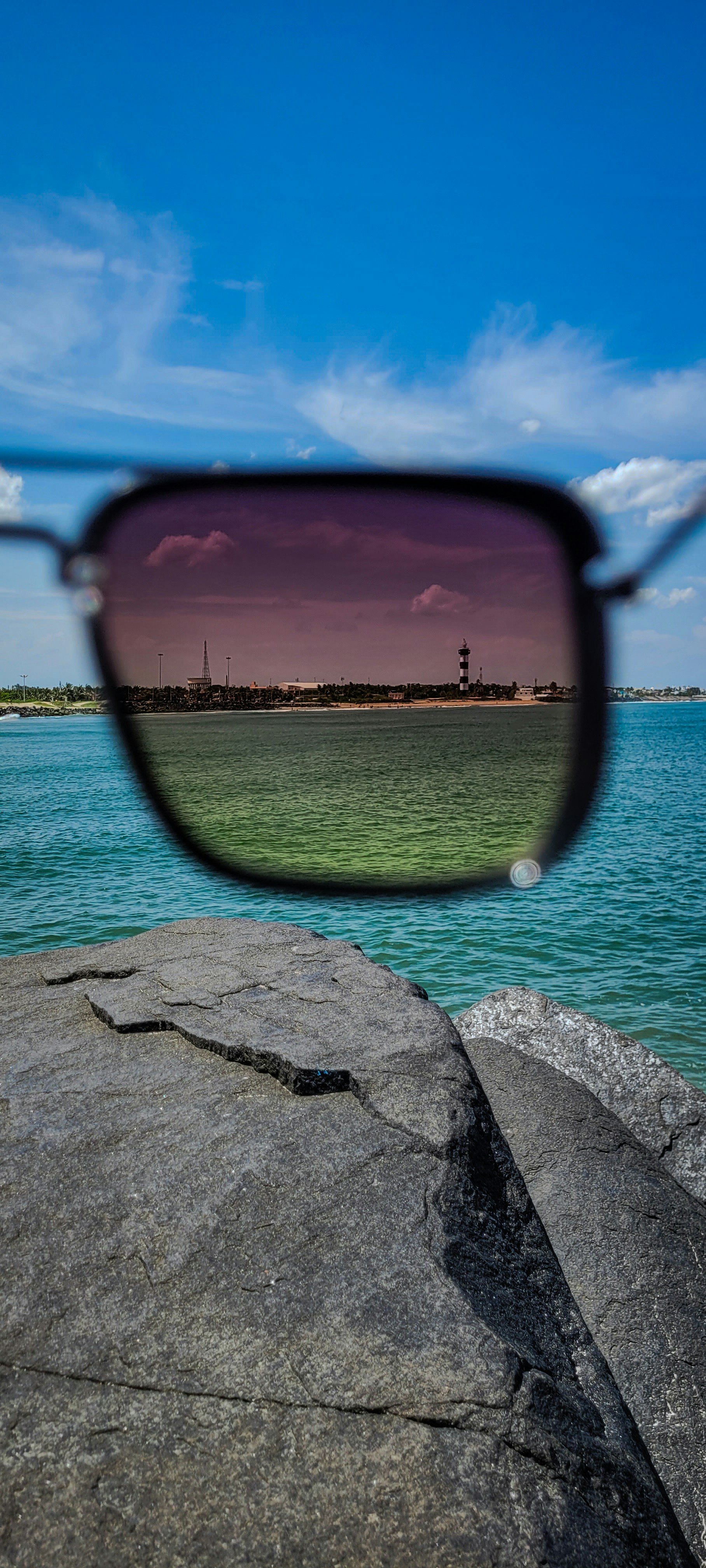 View through a dark sunglass lens: the sea inside the lens looks green and clear, the water around it bright turquoise, with a lighthouse on the shore