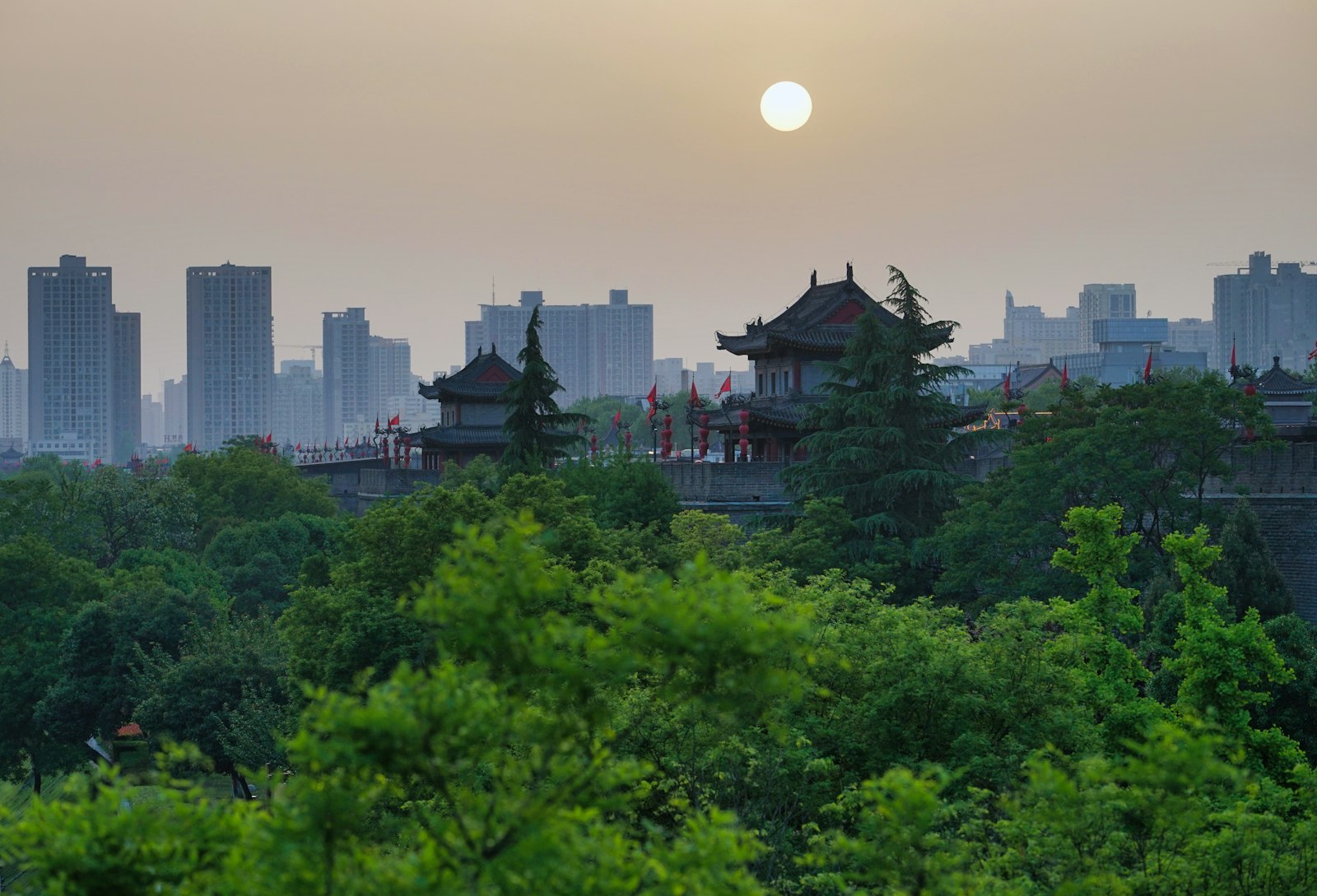 Pagoda-roofed gate towers of Xi'an's city wall rising from trees at hazy sunset, with modern towers on the skyline