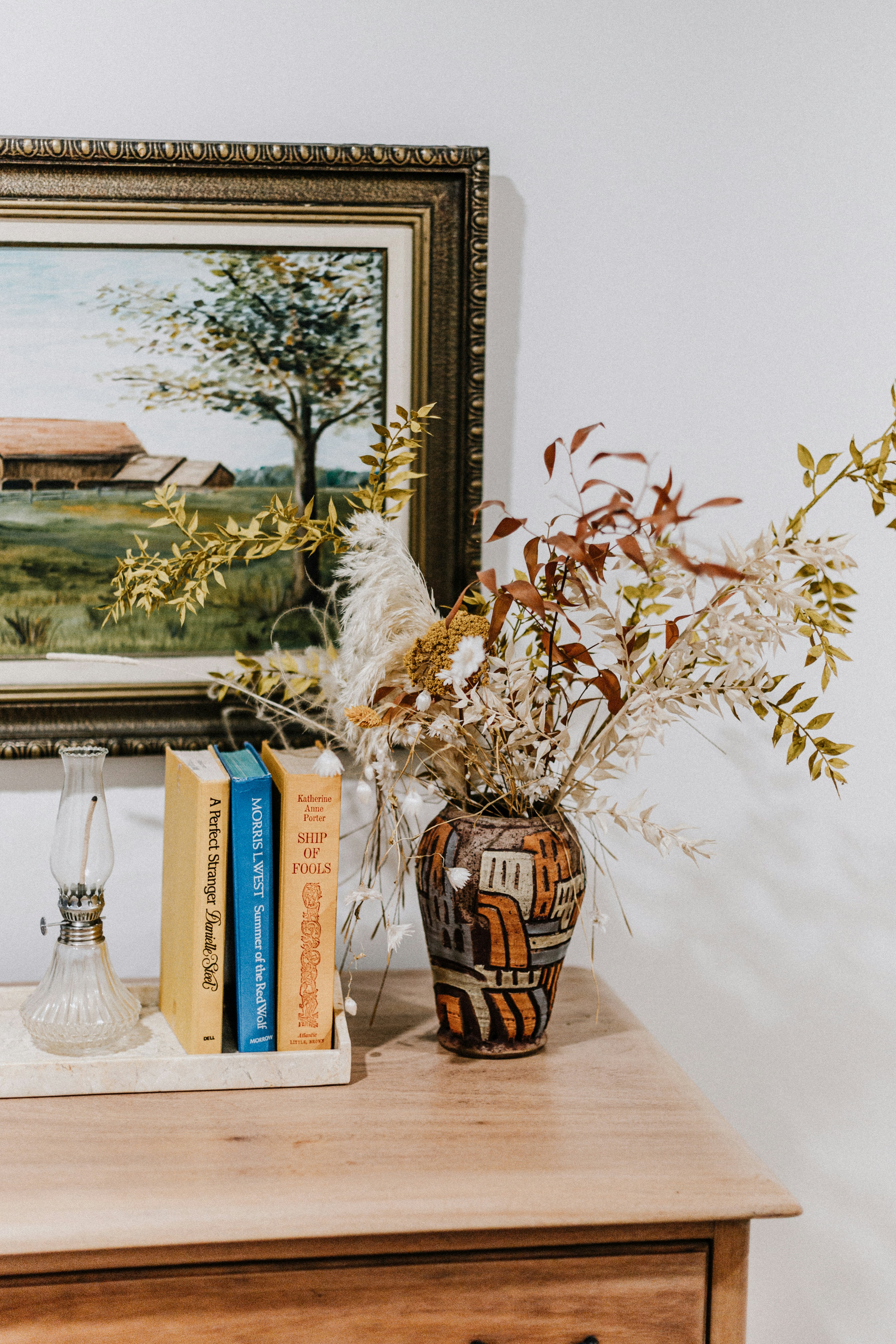 A painting of a farm and a vase of flowers on a dresser