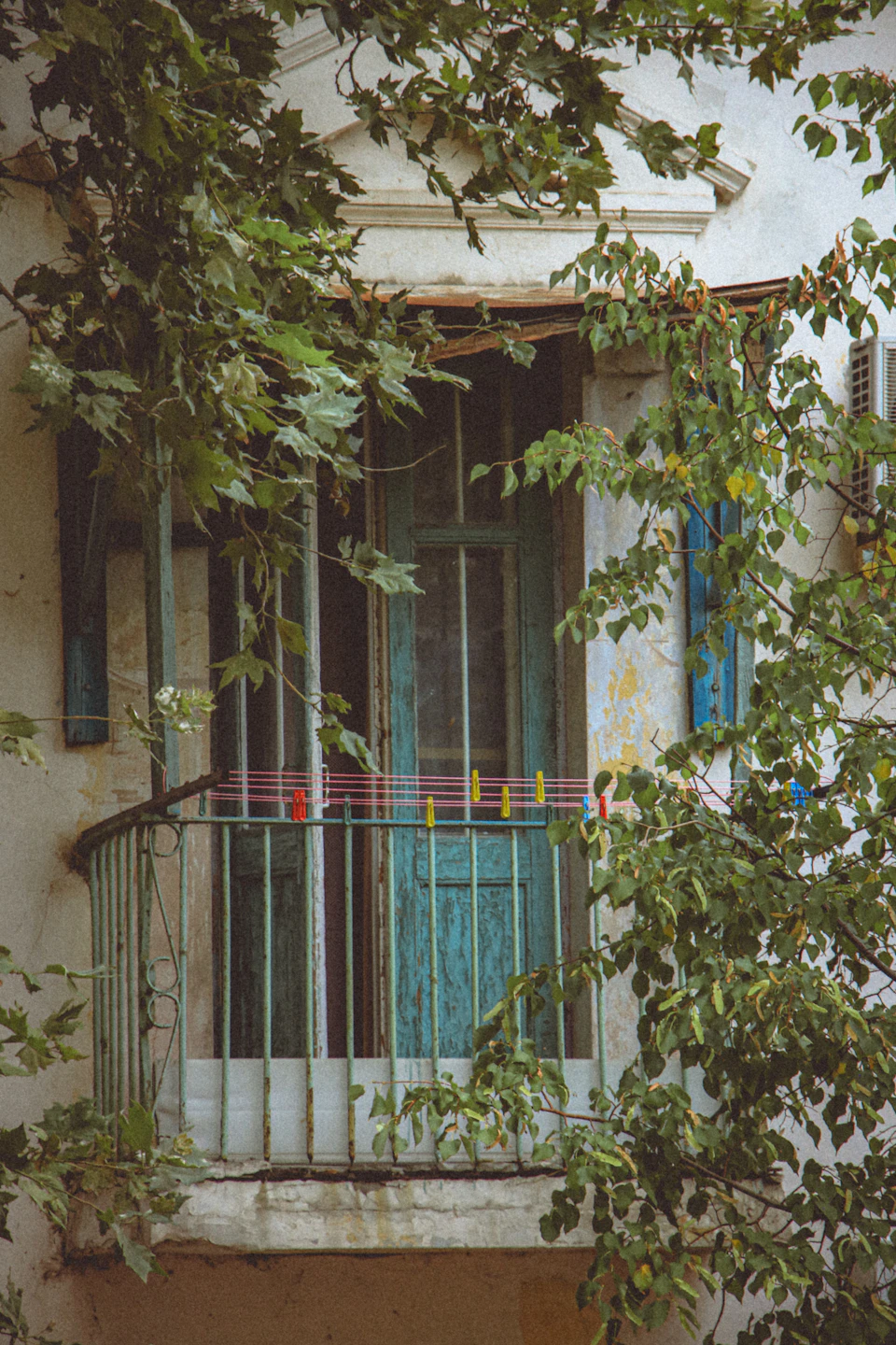 The image shows a quaint balcony with a green railing and a blue door, situated on the second floor of a white building. The balcony is adorned with lush green plants that provide a natural canopy over the entrance. A small window can be seen in the background, adding depth to the scene.