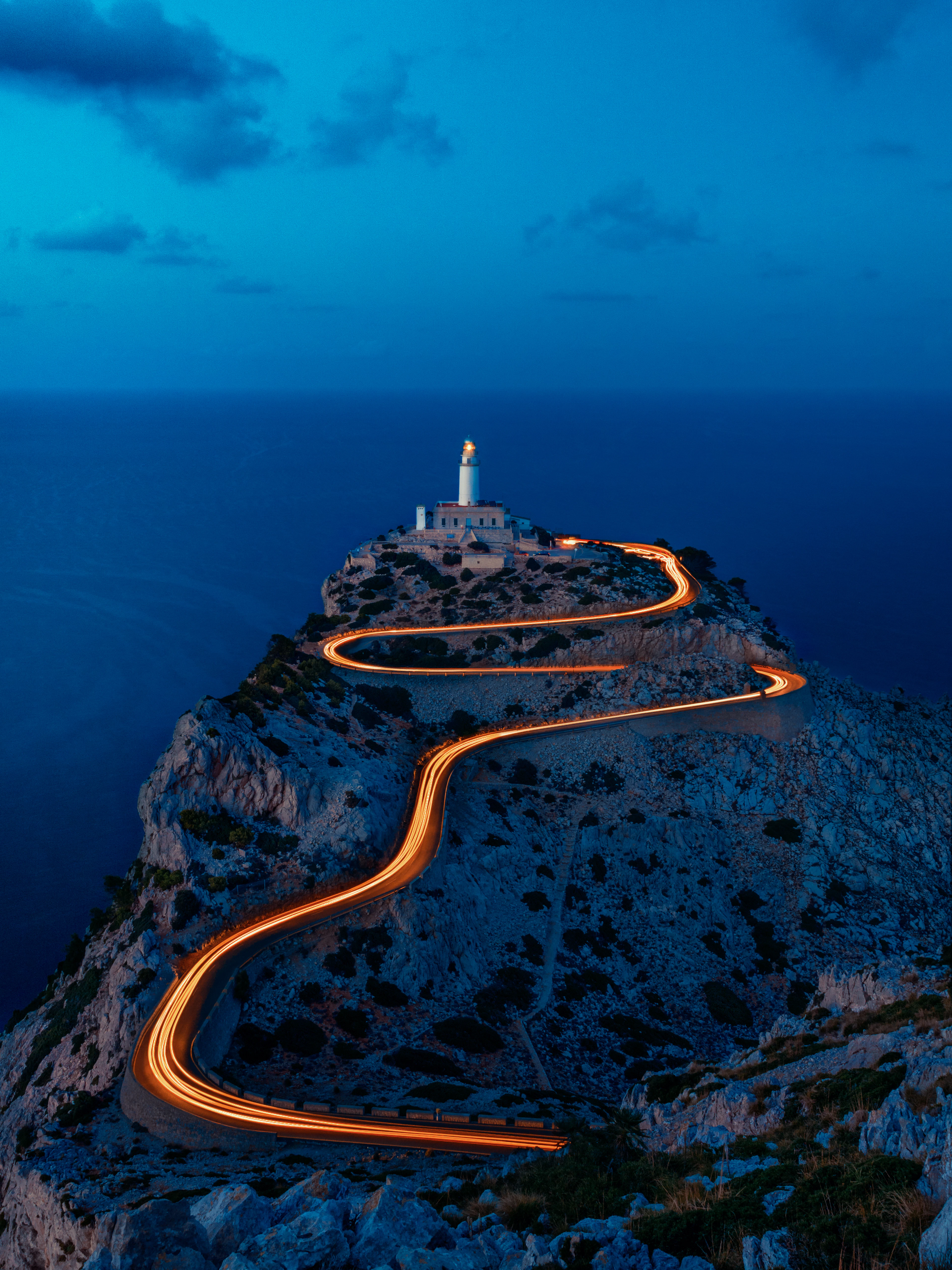 The lighthouse at Cap de Formentor on Mallorca's northern tip at blue hour, the headland road below drawn as a ribbon of orange car-light trails winding up the cliff above a deep blue sea