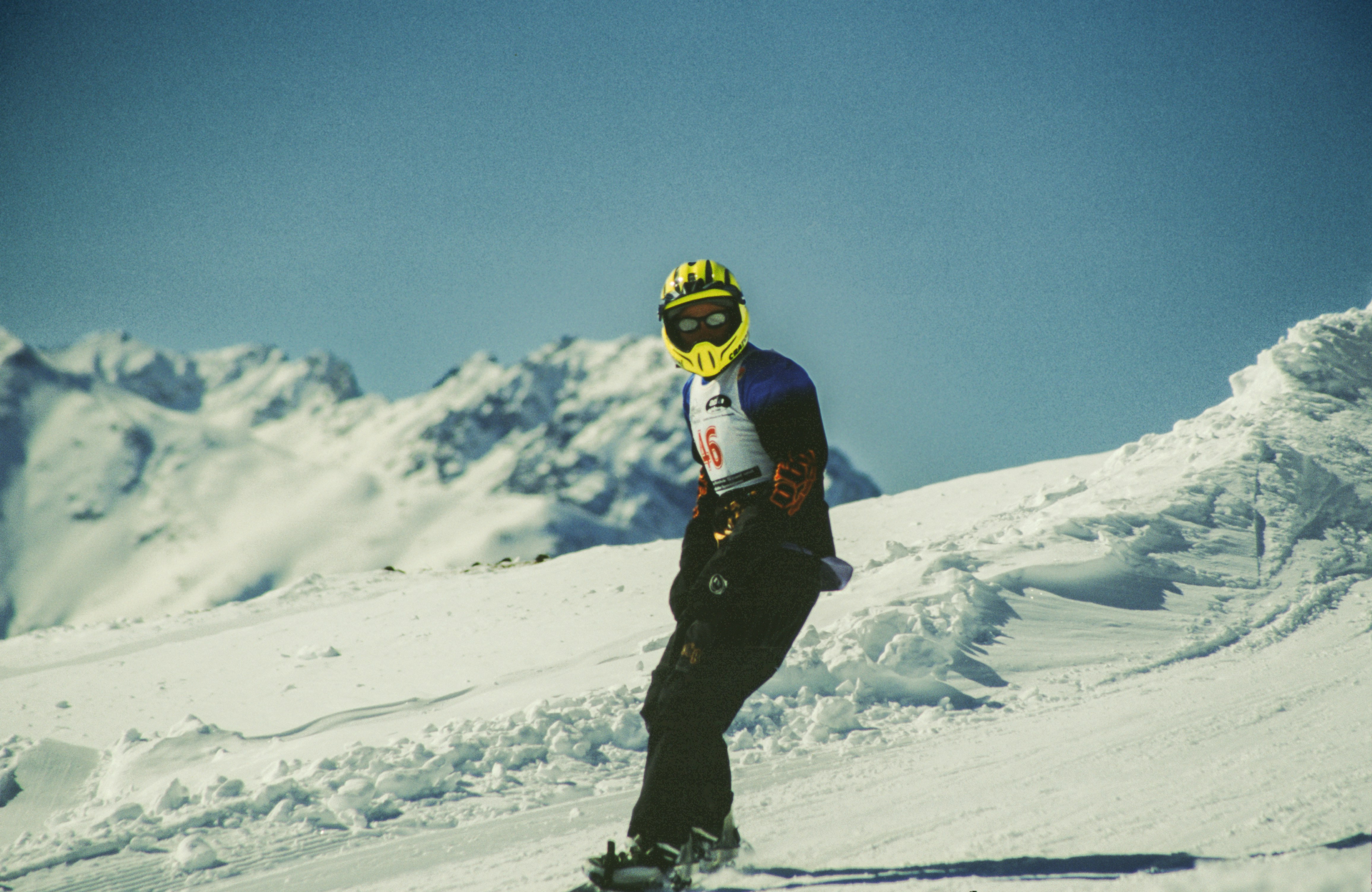 A snowboarder dressed in winter gear, including a bright yellow helmet and goggles, navigates down a snowy slope with snow-covered mountains in the background. The person is wearing a race bib with the number 46, indicating participation in a competition. The scene captures the athlete in motion against a clear blue sky, surrounded by snow and rugged alpine terrain, creating a dynamic winter sports setting. Taken 1998 in Scoul on film