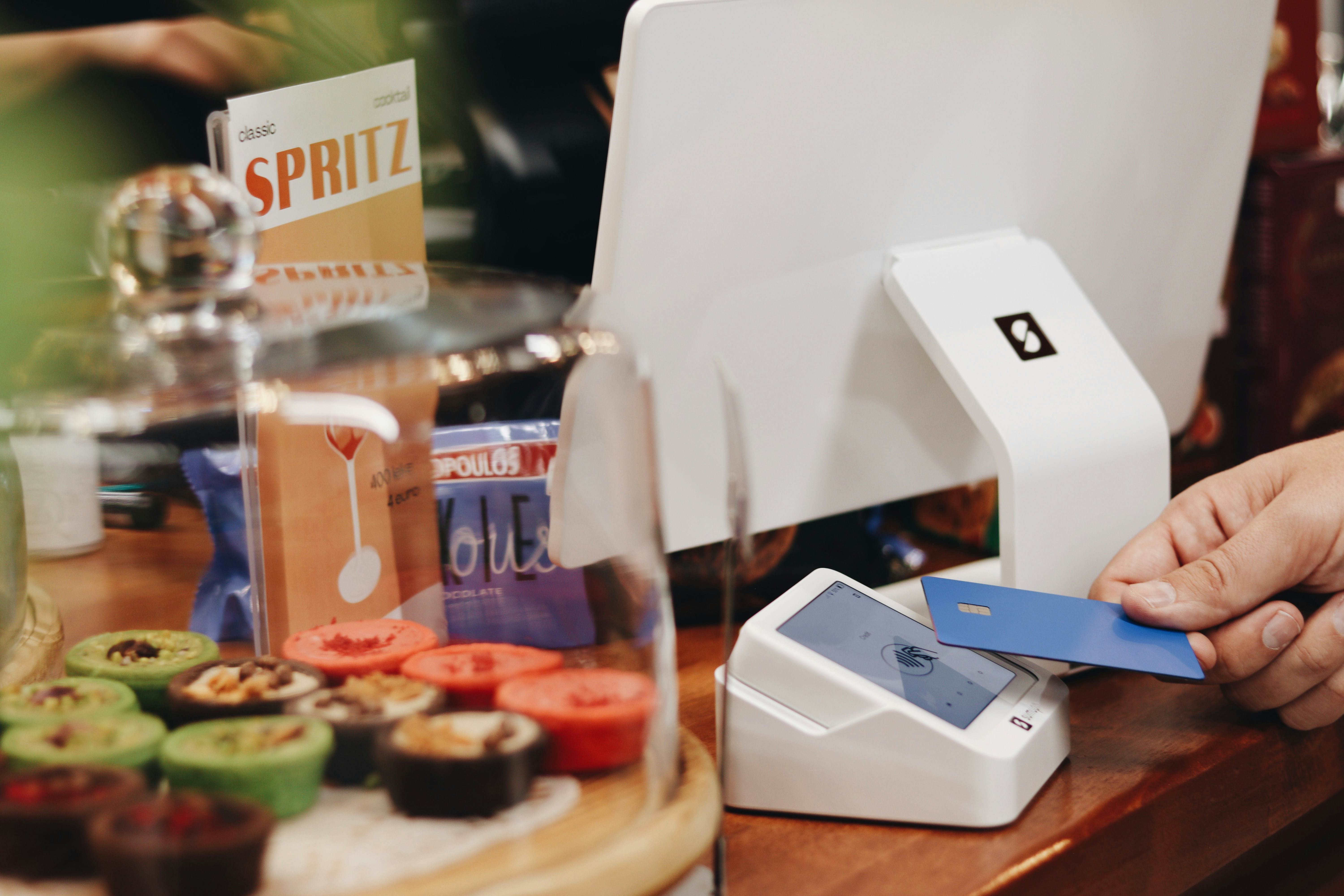 A customer is making a contactless payment using a blue card on a SumUp card reader in a cozy coffee shop or bakery. In the foreground, a variety of colorful desserts are displayed under a glass dome, adding to the inviting atmosphere of the store. A small sign featuring the word \