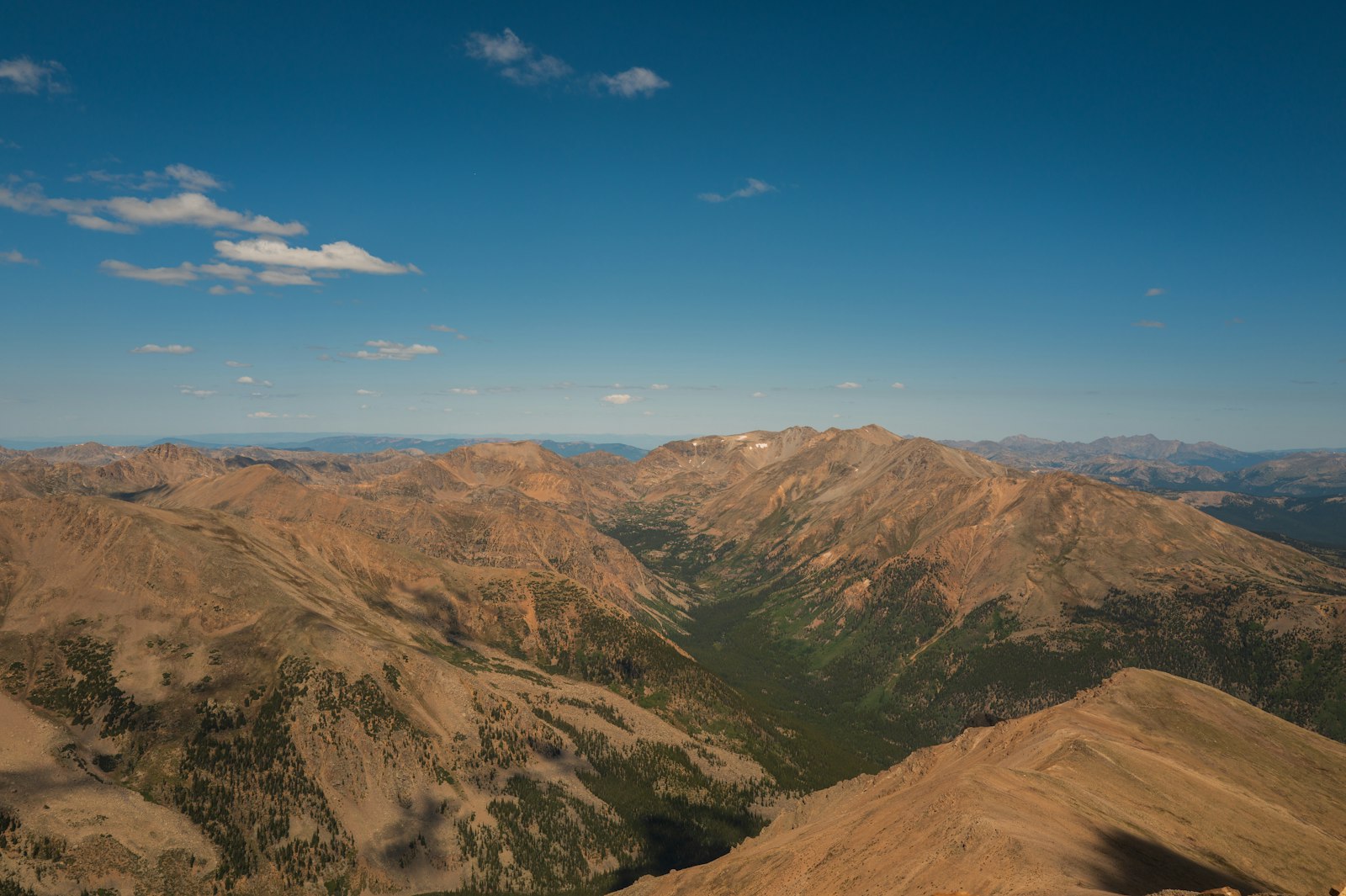 Sunlit brown ridgelines and a forested green valley seen from high on a mountain under a clear blue sky