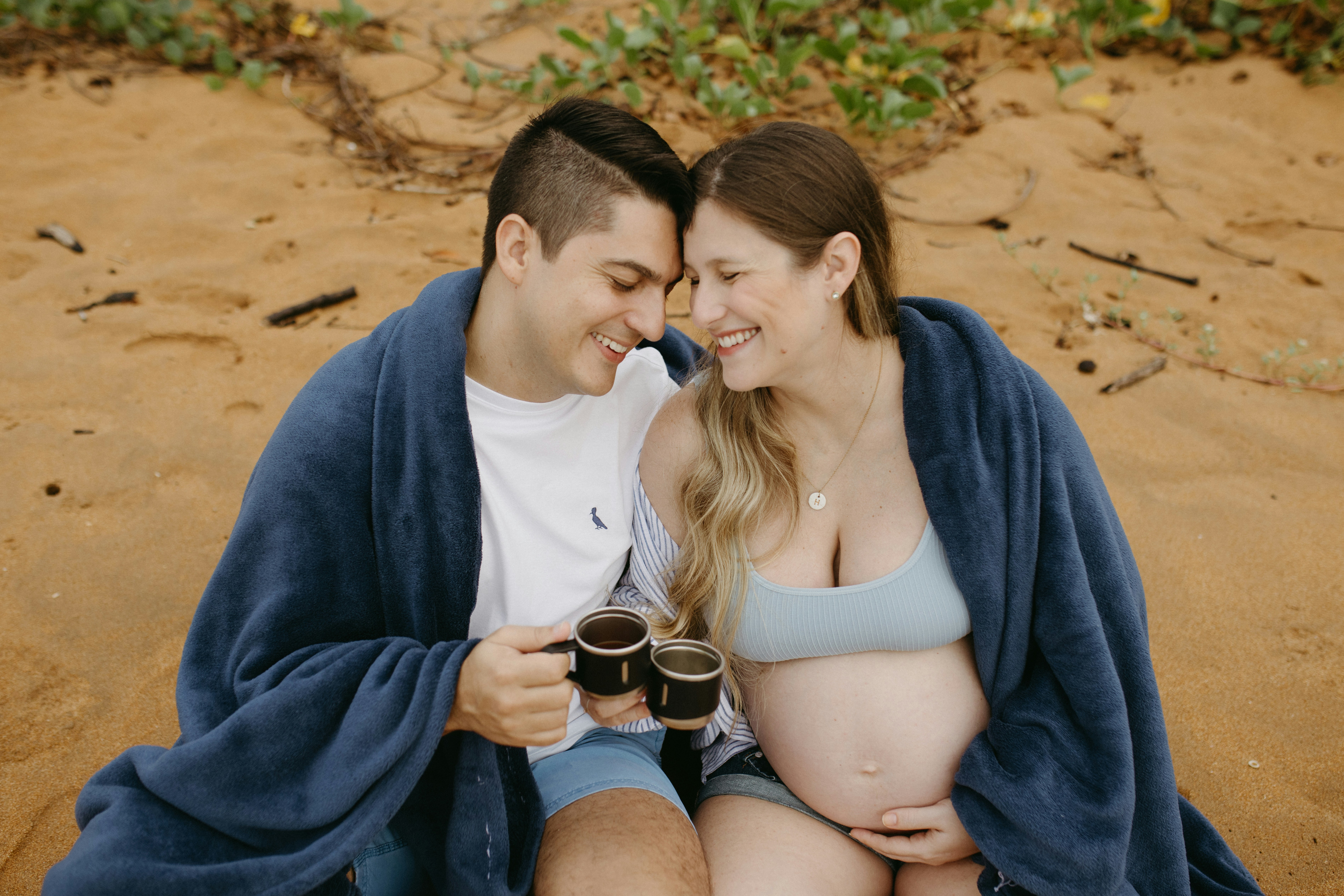 A pregnant couple wrapped in a blanket on the beach
