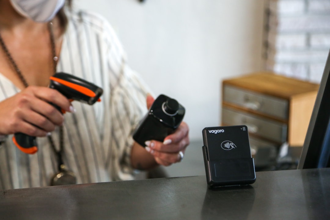 A woman wearing a face mask holding two cameras, A woman at a front desk is scanning a hair styling product with a barcode scanner. The front desk has a credit card reader on it with the Vagaro logo on it.