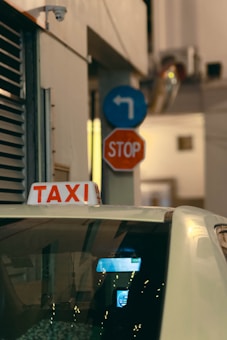 A yellow taxi cab is parked in front of a black SUV, both located near an airport terminal. The taxi displays a phone number and website for Yellow Cab Co. A Hertz rental truck is visible in the background, along with modern architecture and palm trees. The scene is well-lit with a clear blue sky.