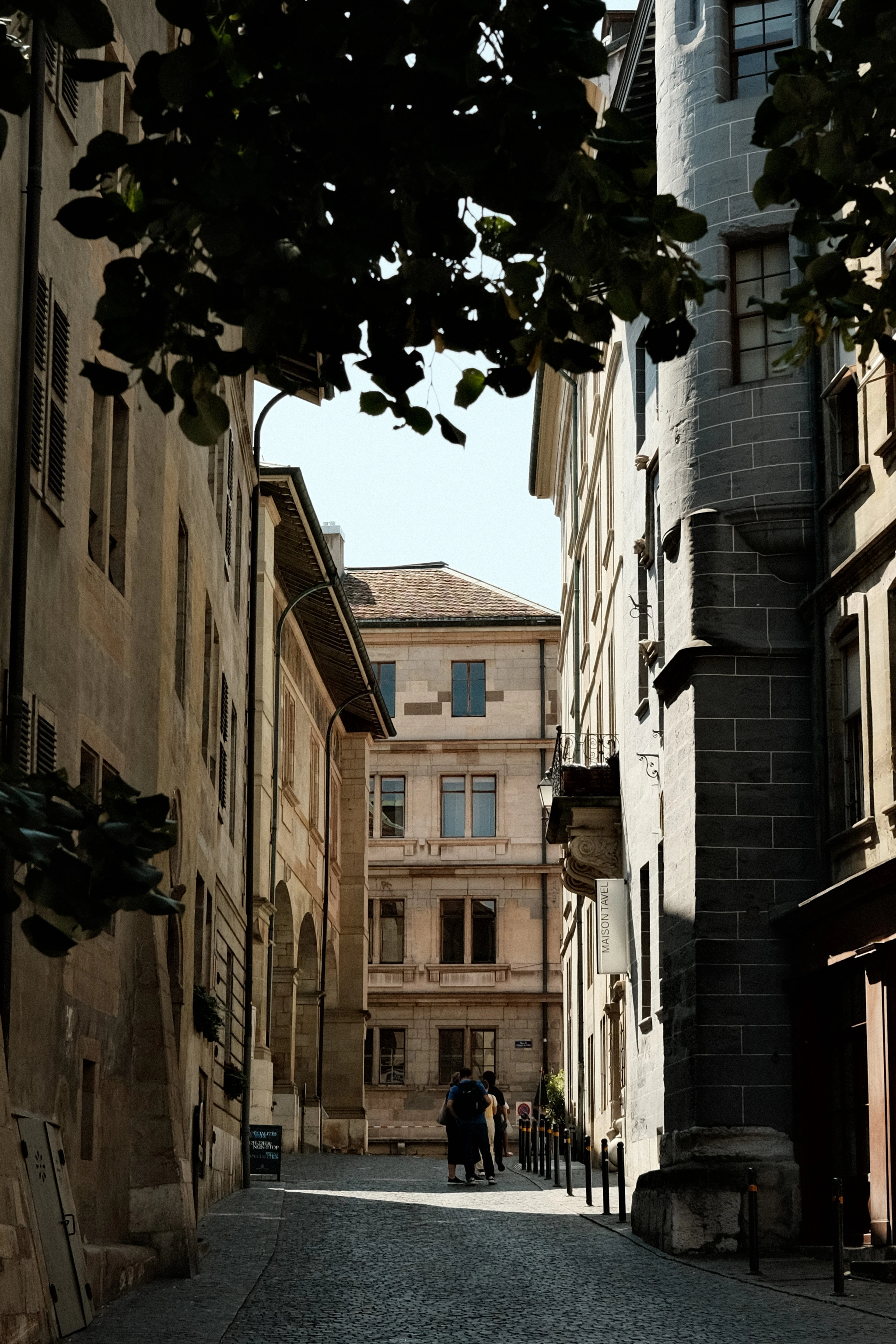 An overhanging tree frames a narrow old-town alley leading to a stone building.