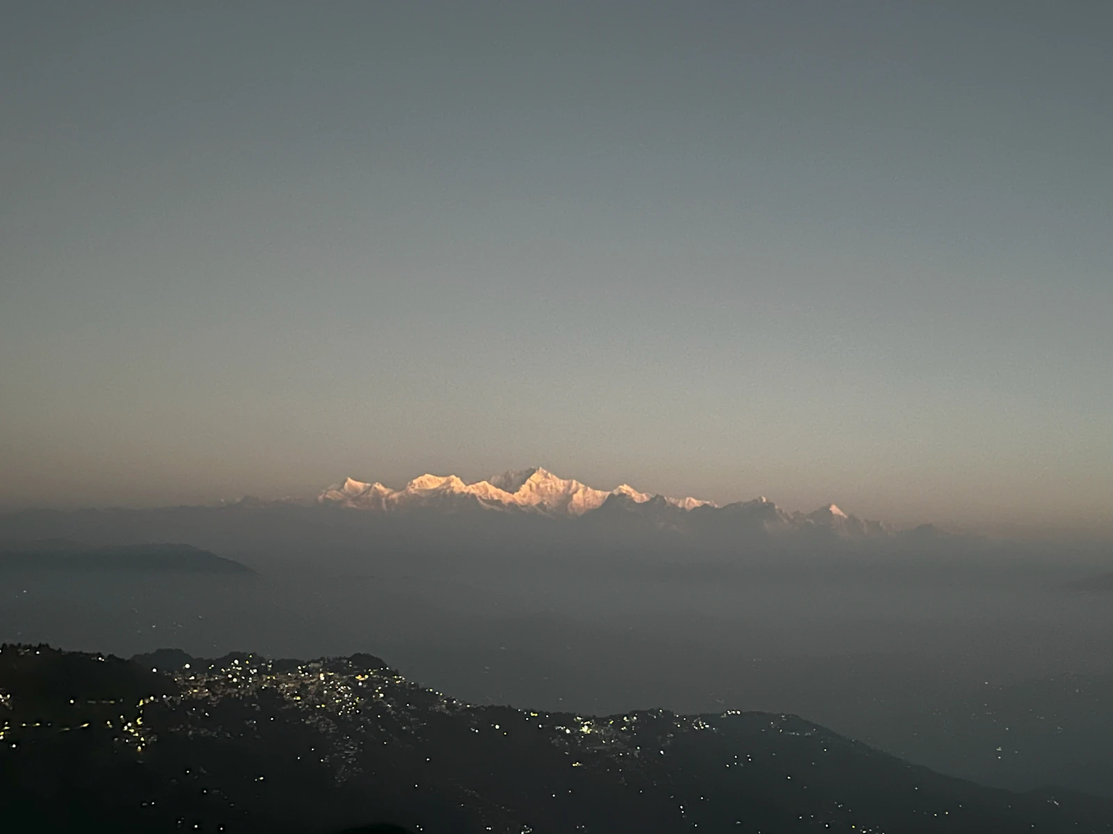 Kanchenjunga lit rose at first light above a haze filled valley, the lights of the hill towns still on below