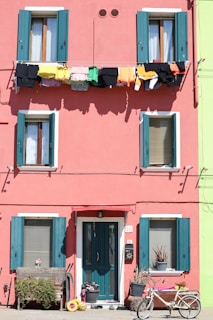 A pink building with green shutters and a bicycle parked in front of it