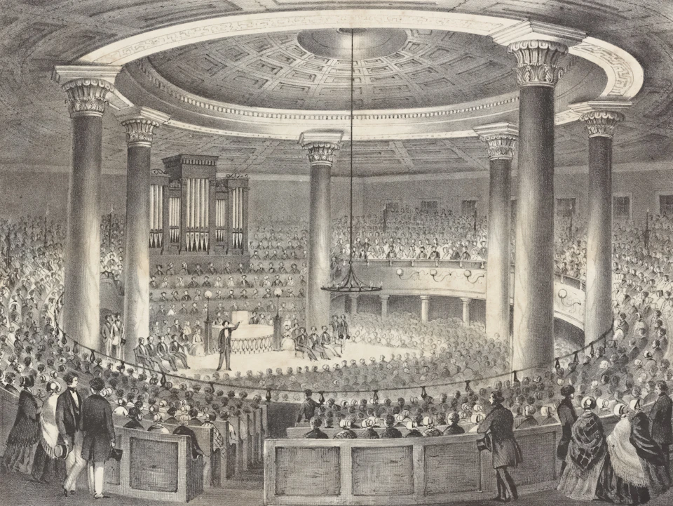 The image depicts a grand church interior with a large circular stage at its center, surrounded by numerous columns and arches that create an imposing architectural structure. The ceiling features a dome-like design, adding to the sense of awe and reverence within the space. A group of people can be seen seated in rows on either side of the stage, their attention focused on the speaker standing before them.