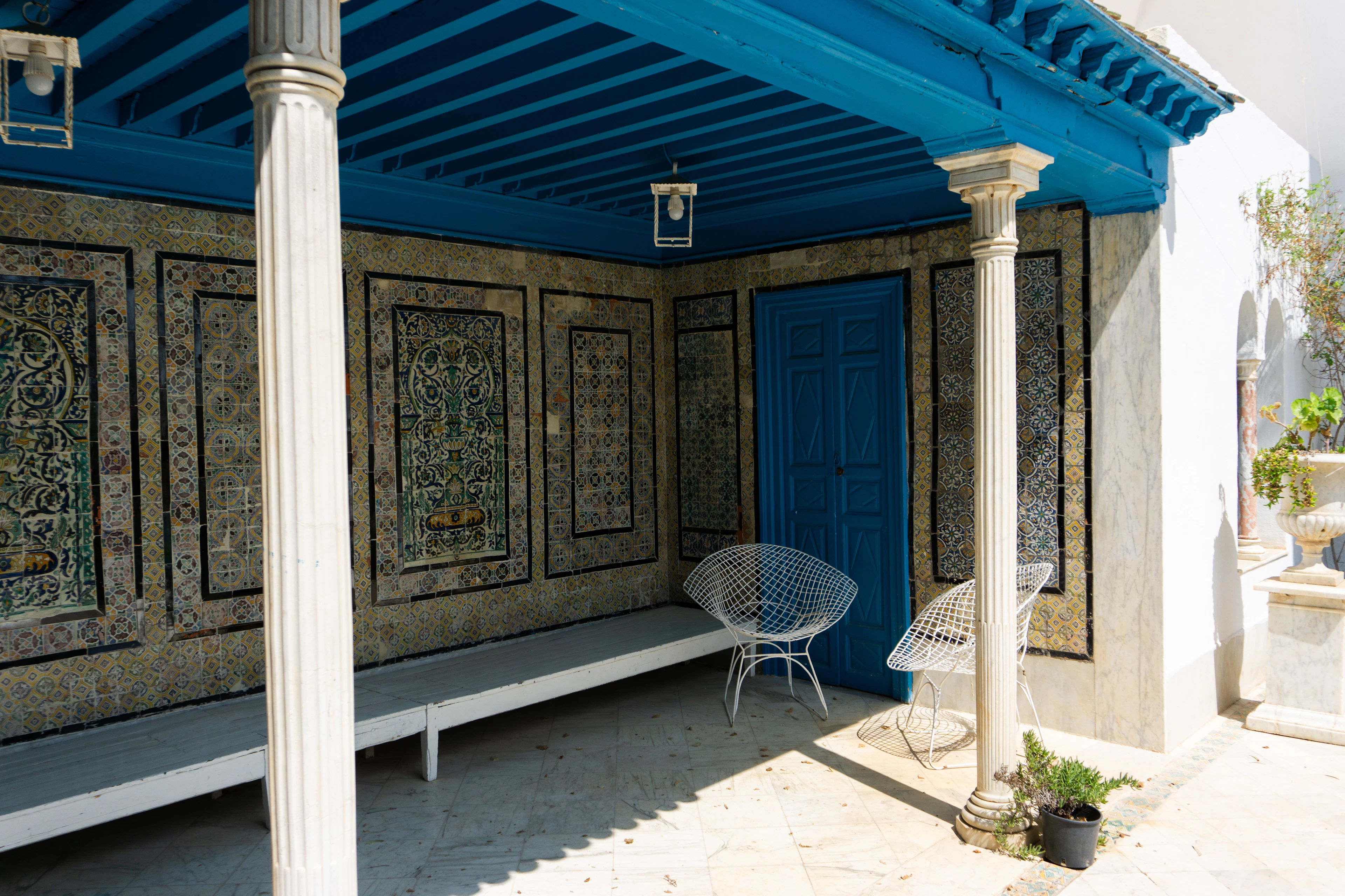 A tiled courtyard portico with a blue beamed ceiling, stone columns, and white wicker chairs.