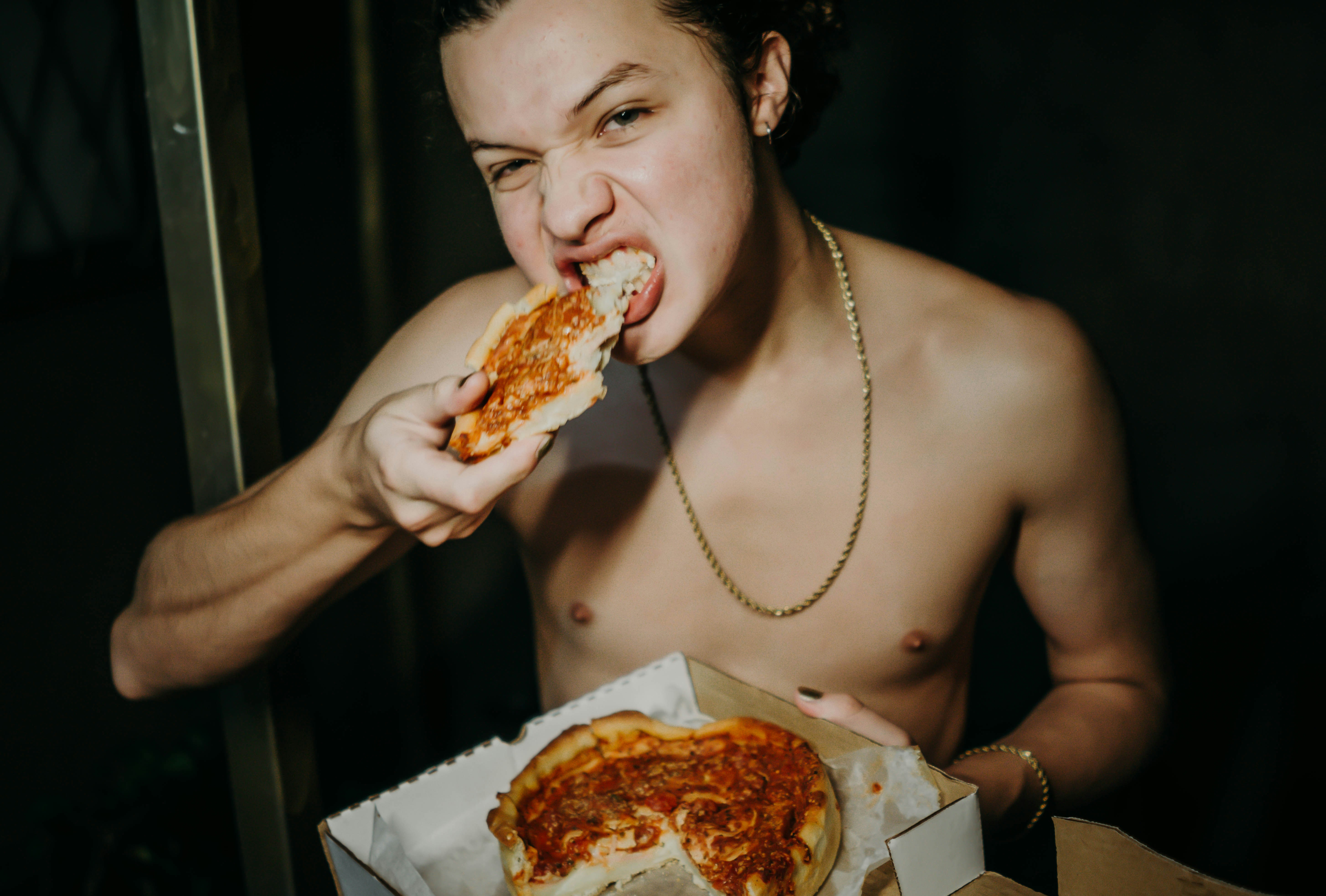 A shirtless man eating a slice of pizza