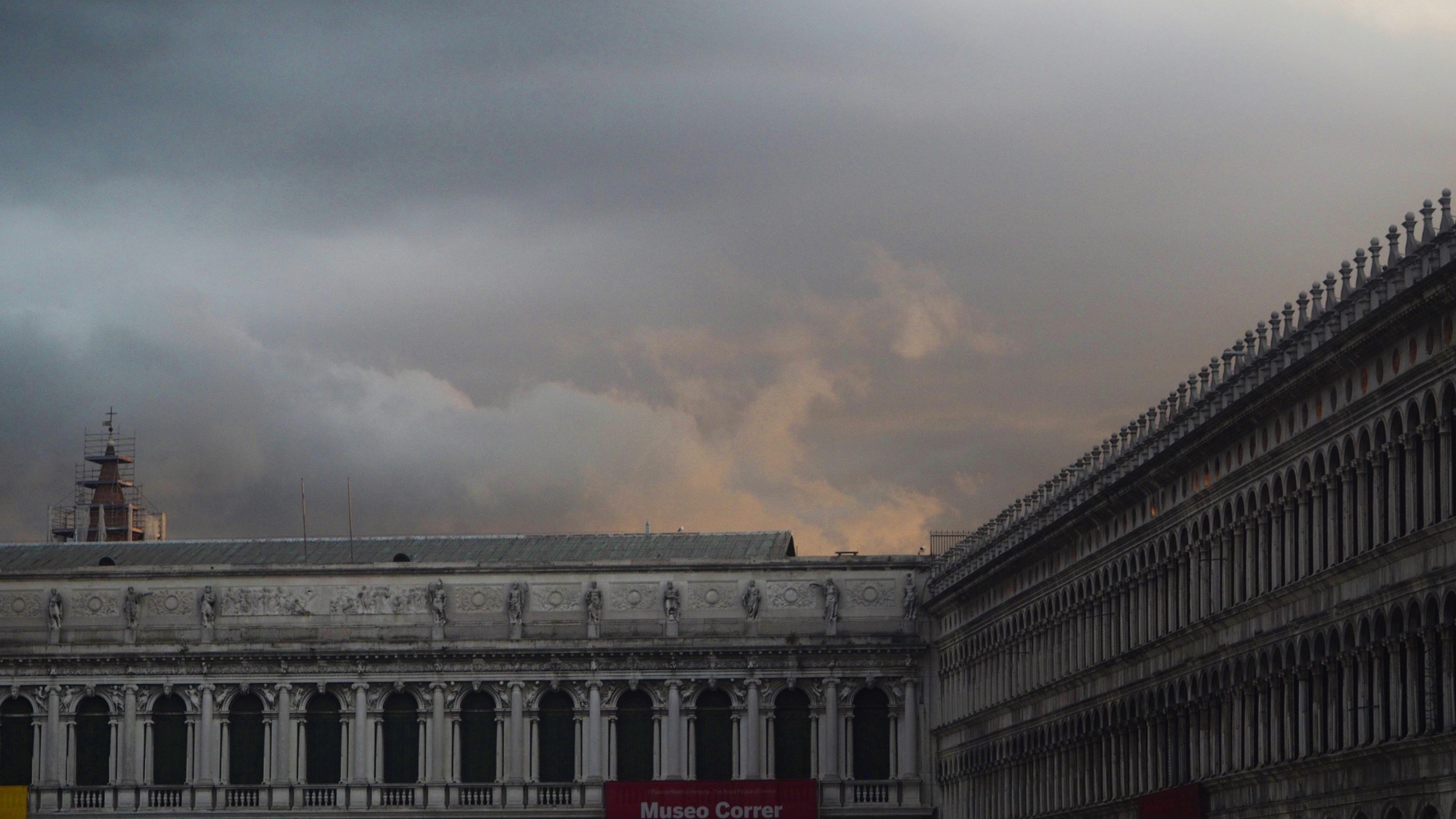 Dramatic view of St. Mark's Square in Venice, looking towards the Museo Correr. The photo captures the grandeur of the historic facades under brooding storm clouds, with rays of sunset light breaking through. The interplay of light and shadow enhances the architectural beauty of the square, showcasing the timeless elegance of Venice against a moody sky.