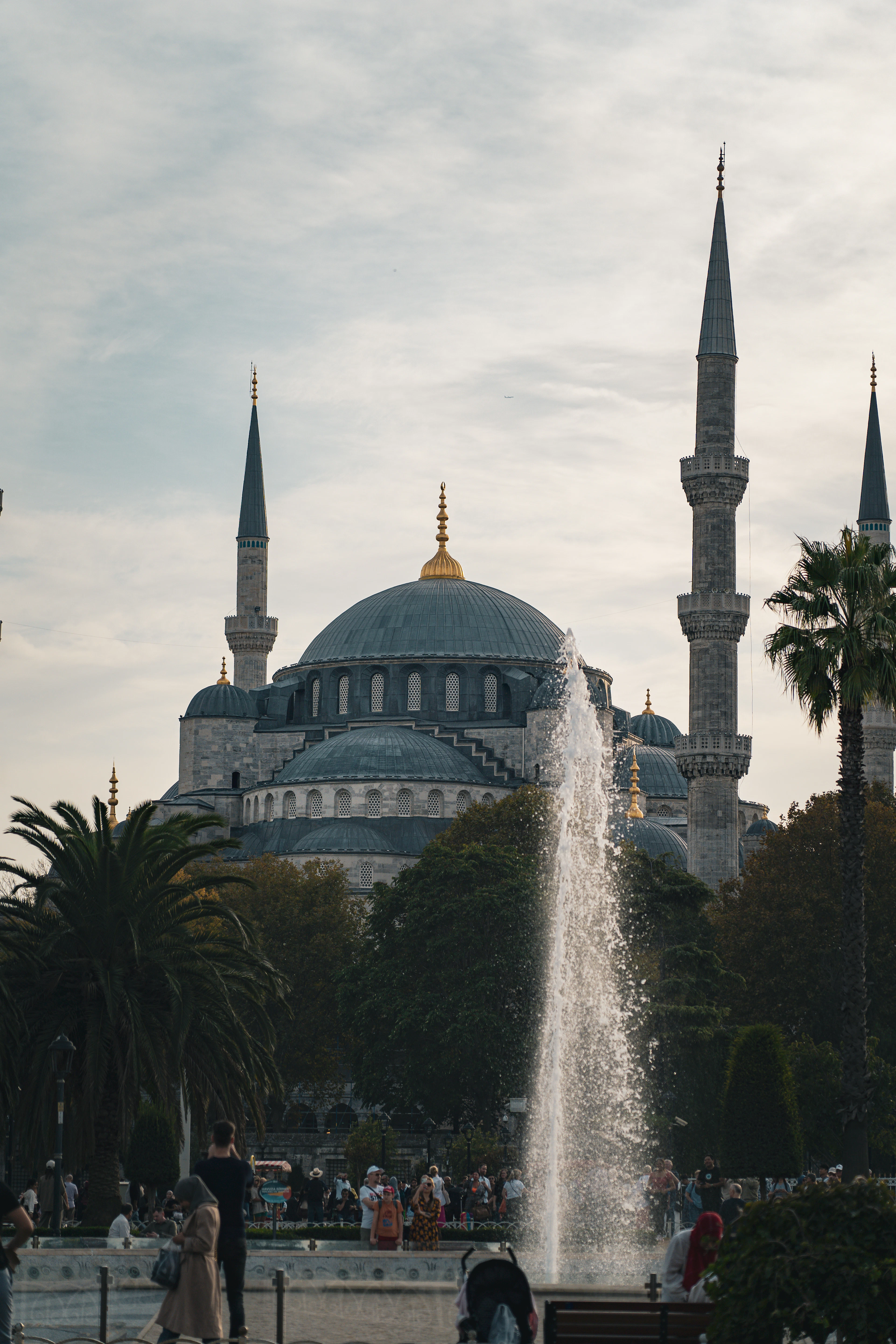 The Blue Mosque's domes rise behind a tall fountain spray at dusk, with palm trees and visitors below.