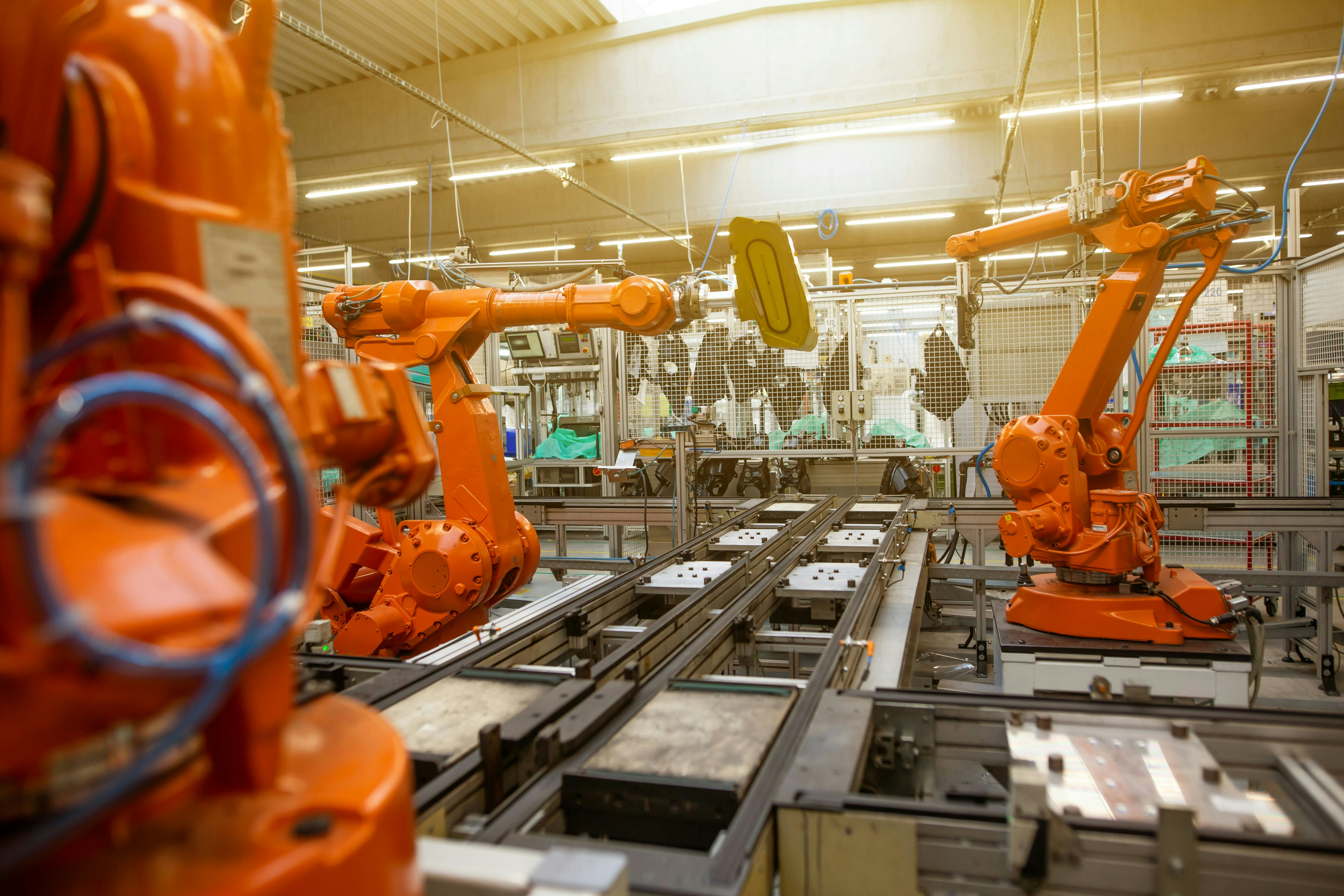 Modern manufacturing facility with robotic assembly arms in well-lit industrial environment, orange safety equipment visible