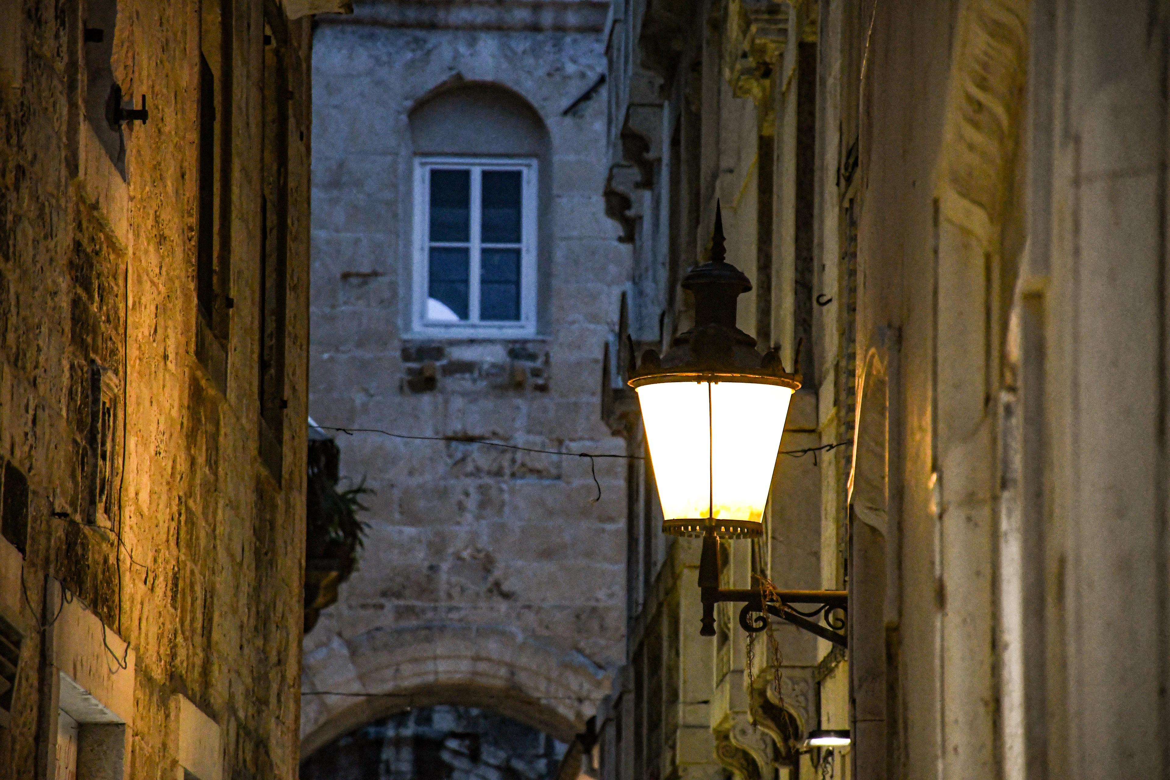 A street lantern glowing at blue hour in a narrow stone lane inside Split's old town, its warm light falling across worn limestone walls and a low arch at the end of the alley.
