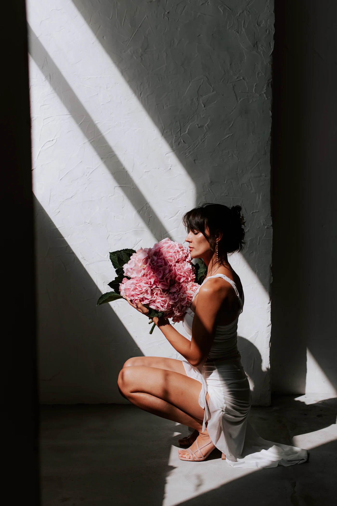 A bride kneels holding a bouquet while a hard diagonal shadow crosses the white wall behind her
