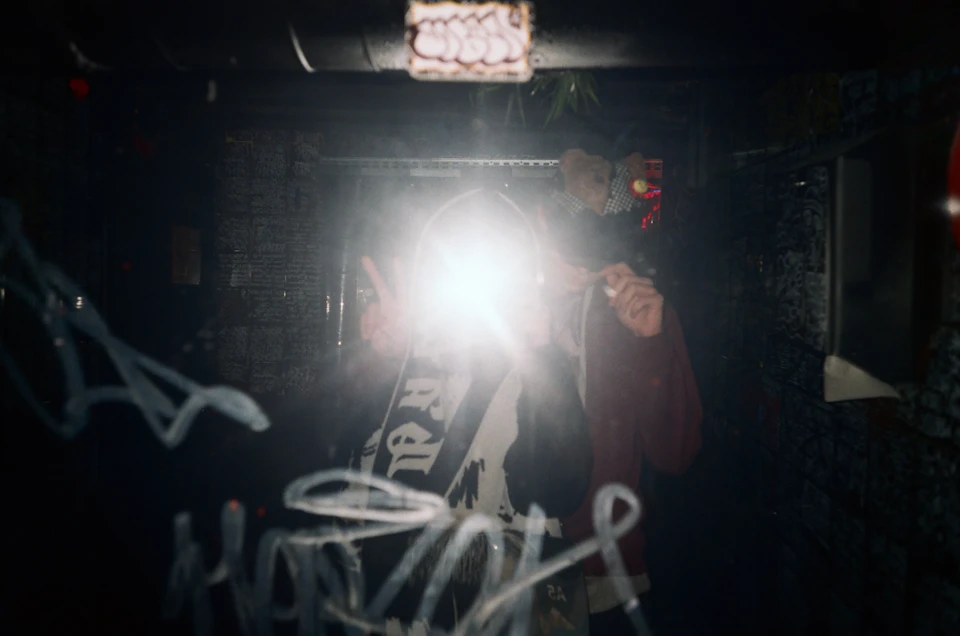 The image shows a dimly lit room with two people standing close together, both holding up their hands in peace signs. The person on the left is wearing a black shirt and has a white sign with black text that reads "MAD". On the right side of the frame, another individual is wearing a red shirt and also holds up their hand in a peace sign.

The room appears to be a basement or underground space, as evidenced by the presence of a brick wall on one side and a window on the other. The overall atmosphere suggests an intimate gathering or event taking place within this enclosed area.